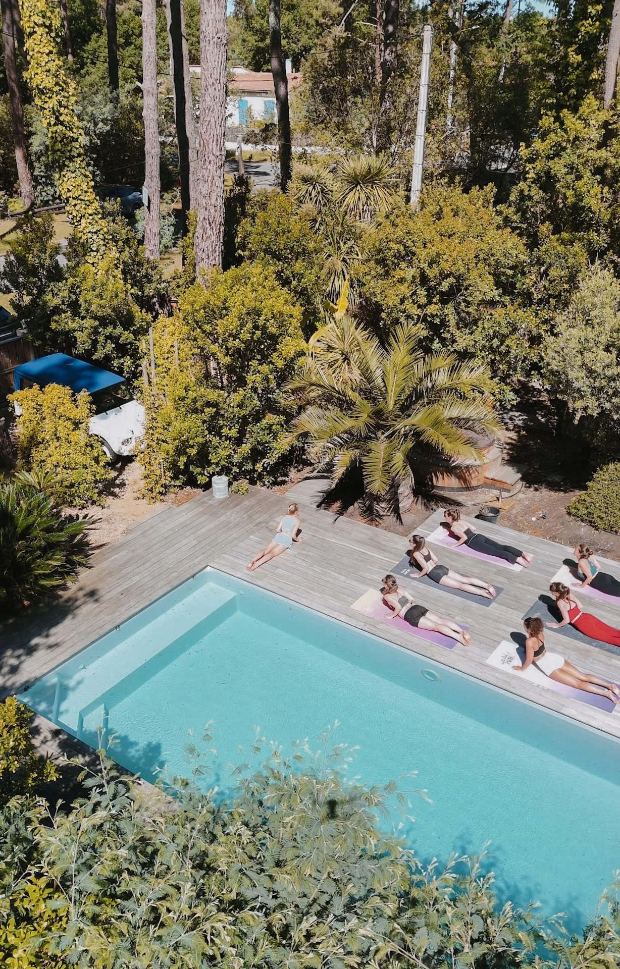 Vue aérienne d'un cours de yoga collectif au bord de la piscine, entourée de pins — surf camp yoga immersif dans la nature landaise