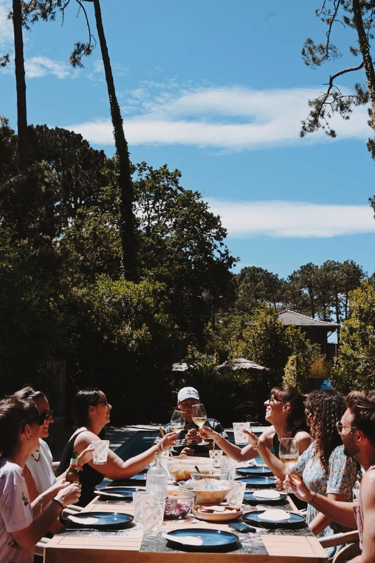 Participants trinquant autour d'une table dressée en terrasse, ombragée par les pins — repas convivial d'une retraite yoga et surf au Cap Ferret