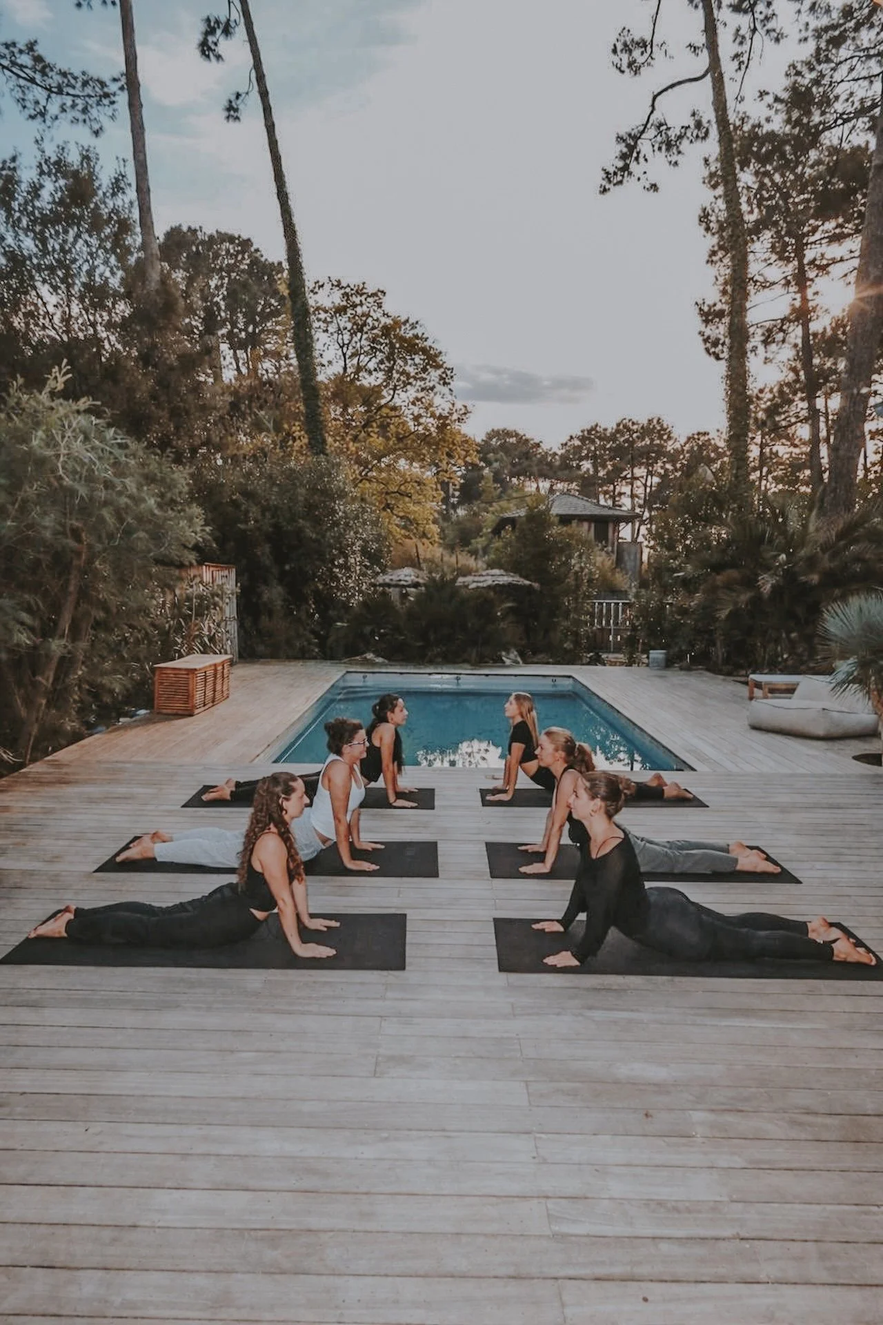 Groupe de participantes en posture du chien tête en haut sur la terrasse en bois, face à face autour de la piscine entourée de pins — cours de yoga collectif lors d'une retraite Cap Ferret