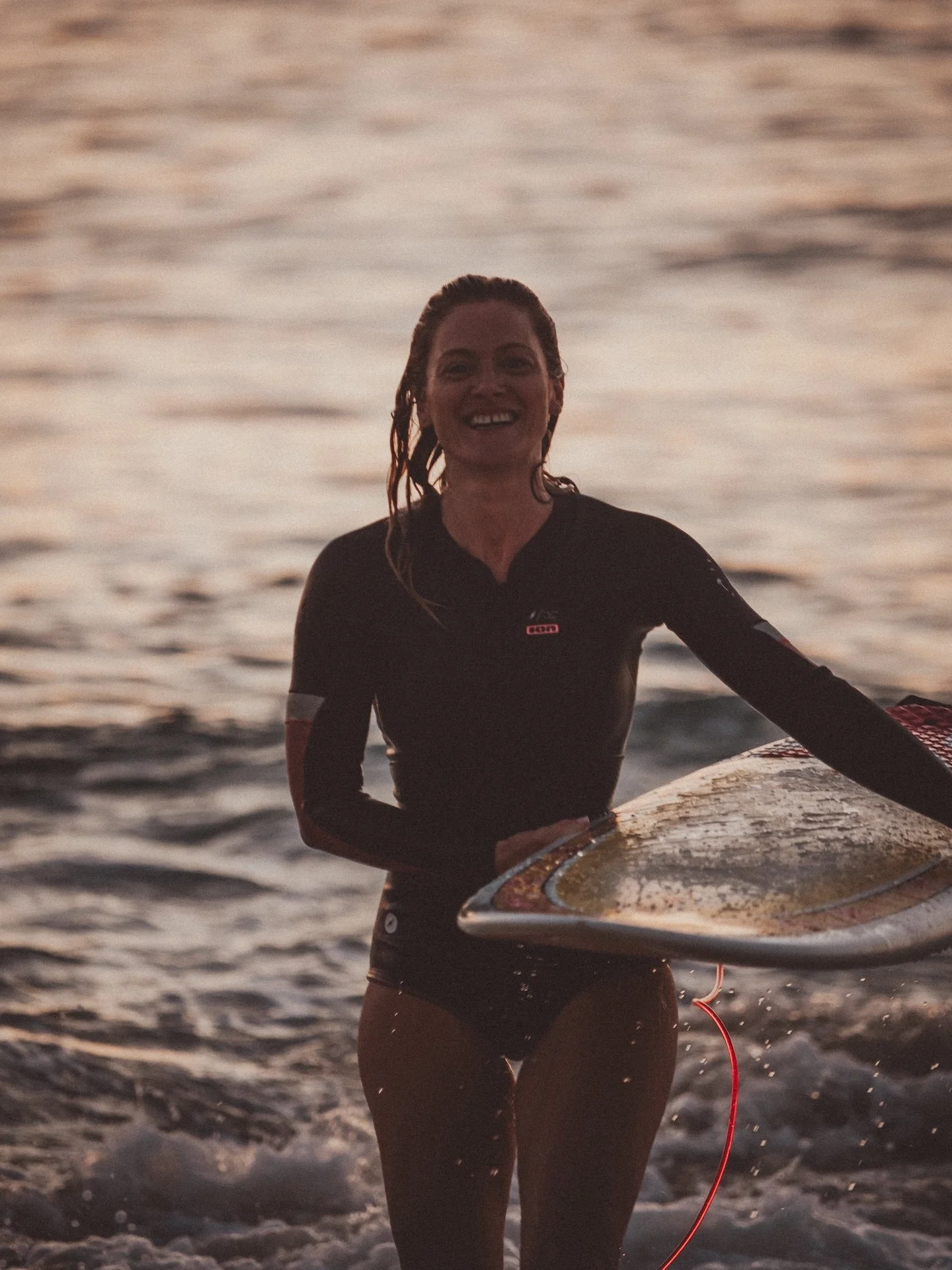 Surfeuse souriante émergeant de l'eau, planche sous le bras, lumière dorée du soir — l'énergie d'une retraite yoga et surf au Cap Ferret