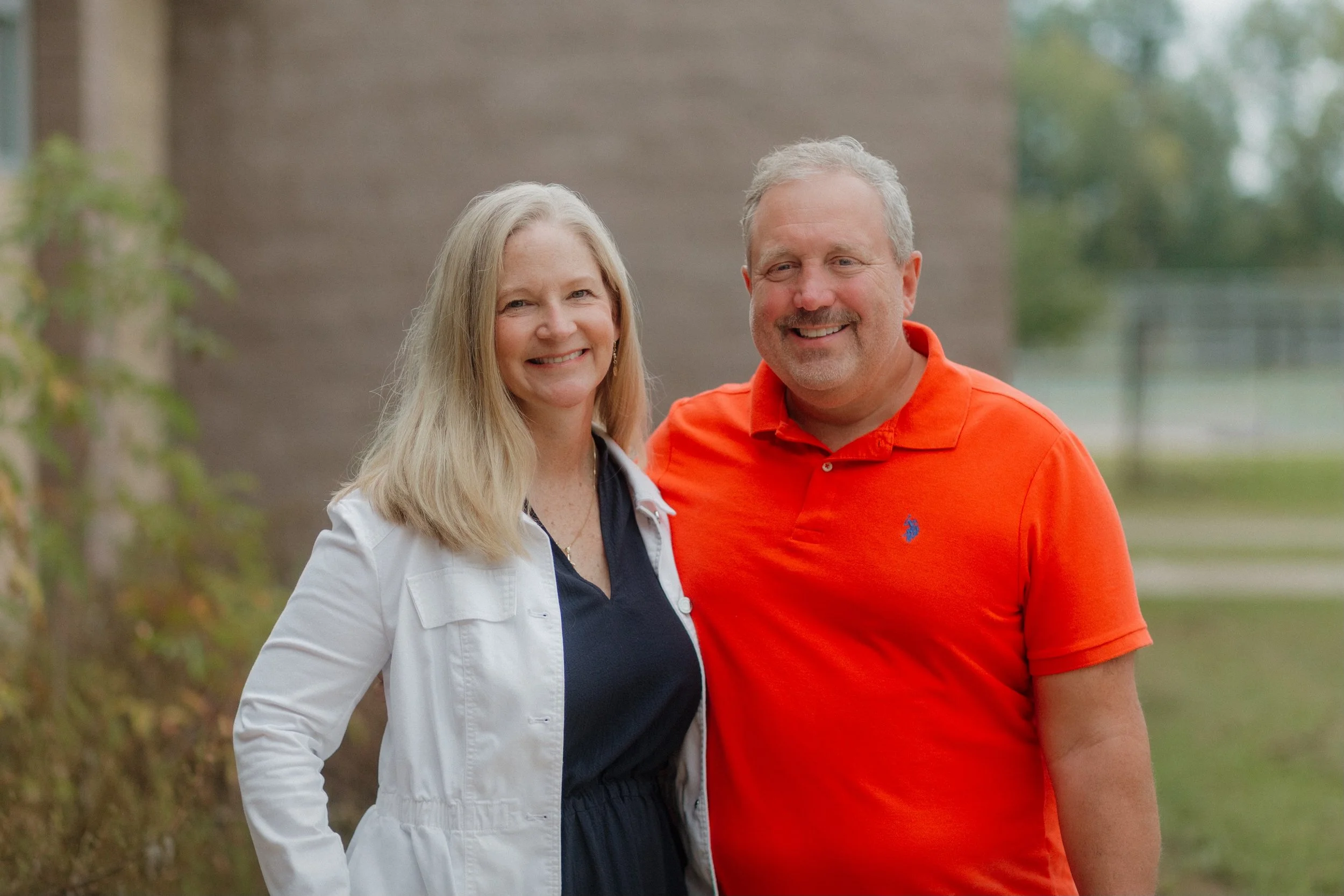 A smiling man and woman taking a selfie outdoors with rolling hills and green fields in the background.