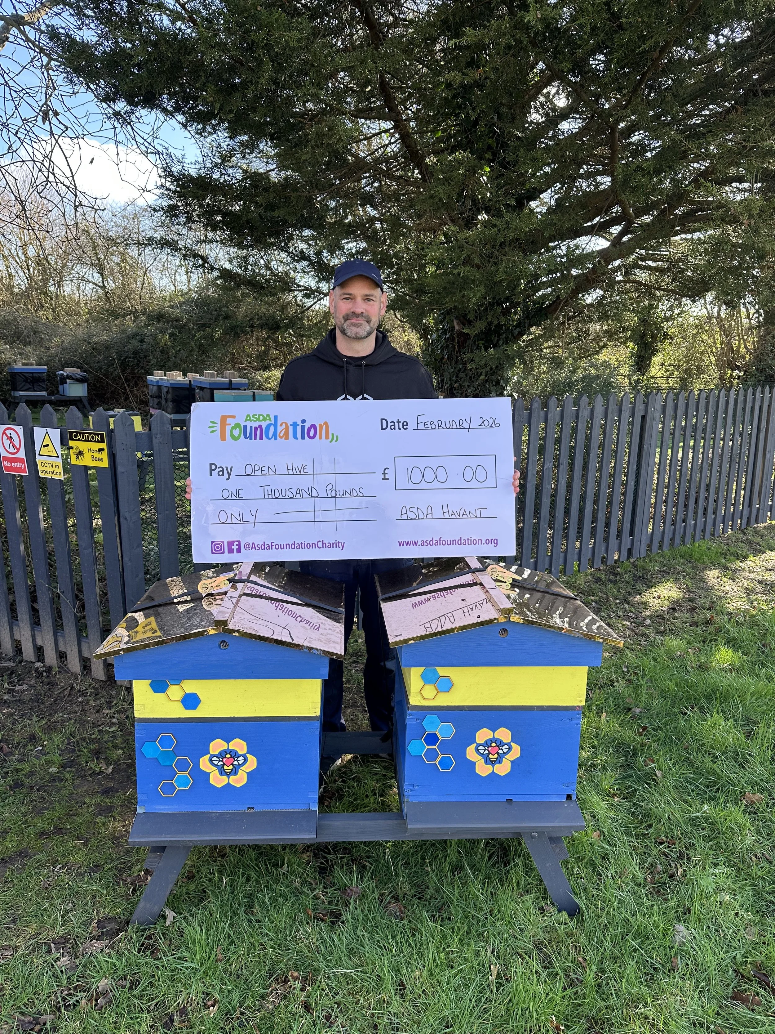 A man standing outdoors holding a large check made out to 'Open Hive' for 1000 pounds, dated February 2023, from ASDA Foundation. He is standing behind two colorful bee-themed beehives painted blue, yellow, and decorated with honeycomb patterns and flowers, near a gray fence and trees.