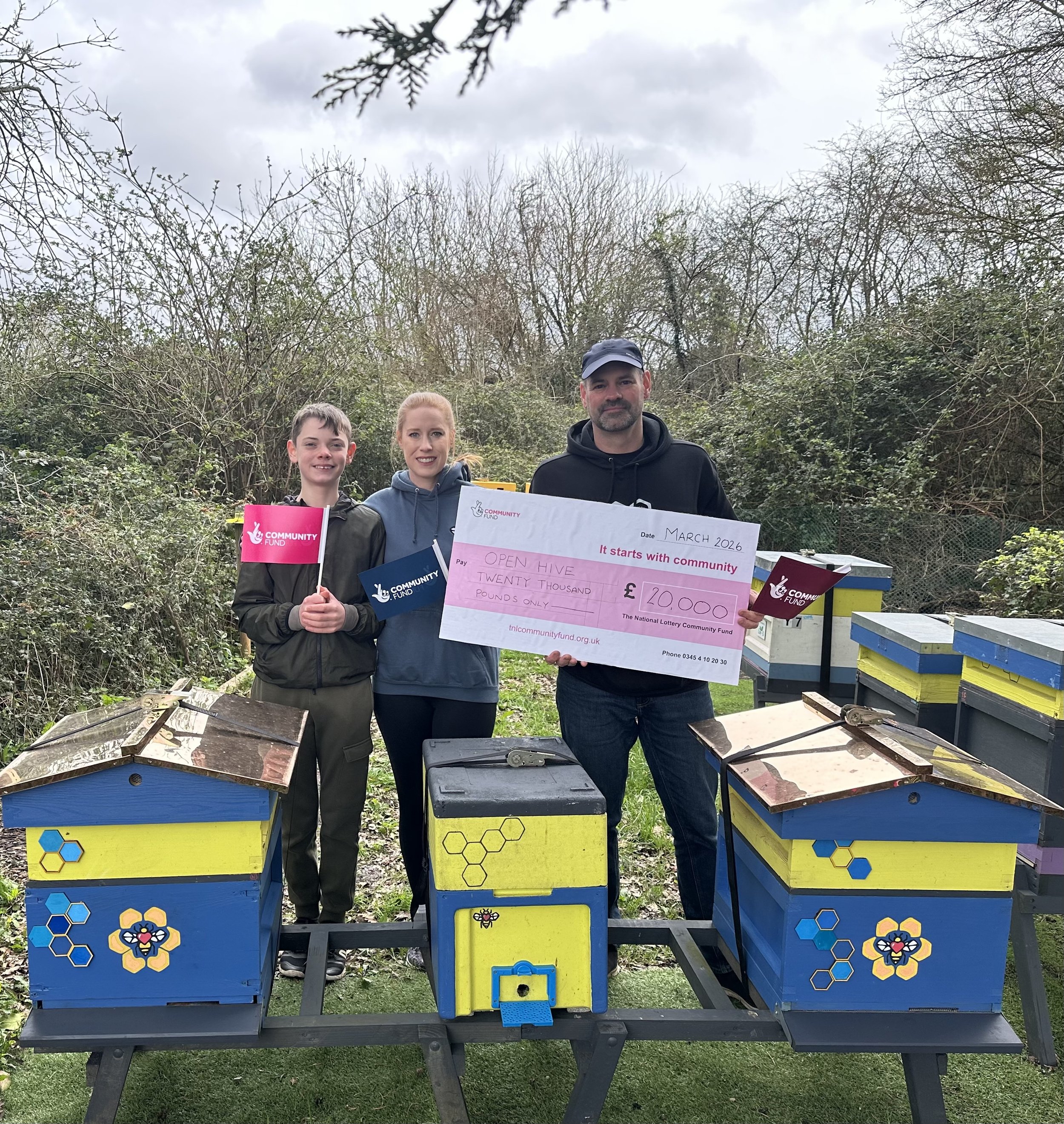 Three people standing outdoors near beehives, holding a large ceremonial check for twenty thousand pounds. The beehives are colorful with honeycomb and bee decorations. One person holds a pink flag, another holds a blue flag, and the third person holds the check.