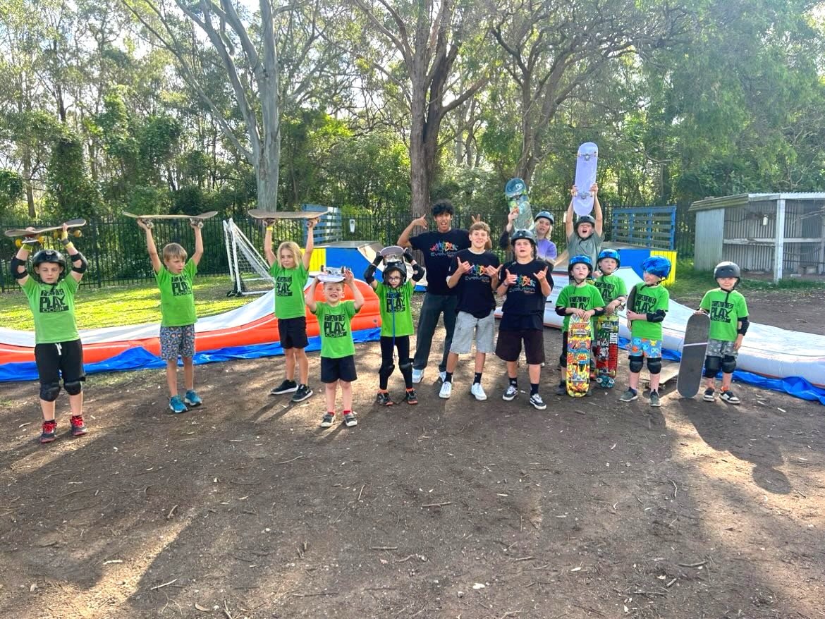 Students and coaches at a skateboard park holding skateboards, some wearing helmets, with a large inflatable ramp in the background.