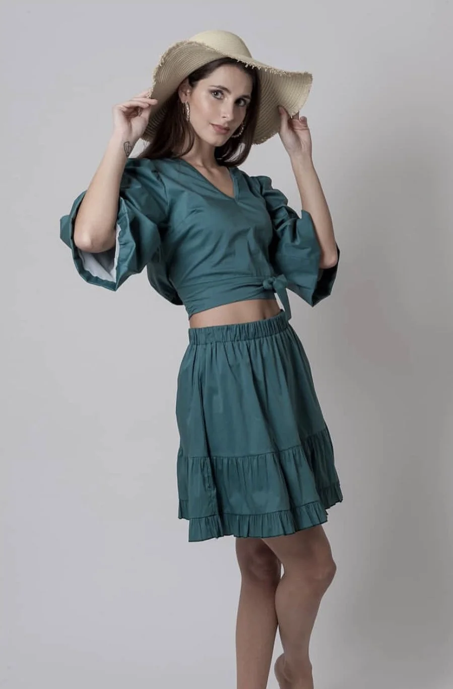 Woman wearing a teal crop top and skirt with a wide-brimmed beige hat, posing against a plain background.
