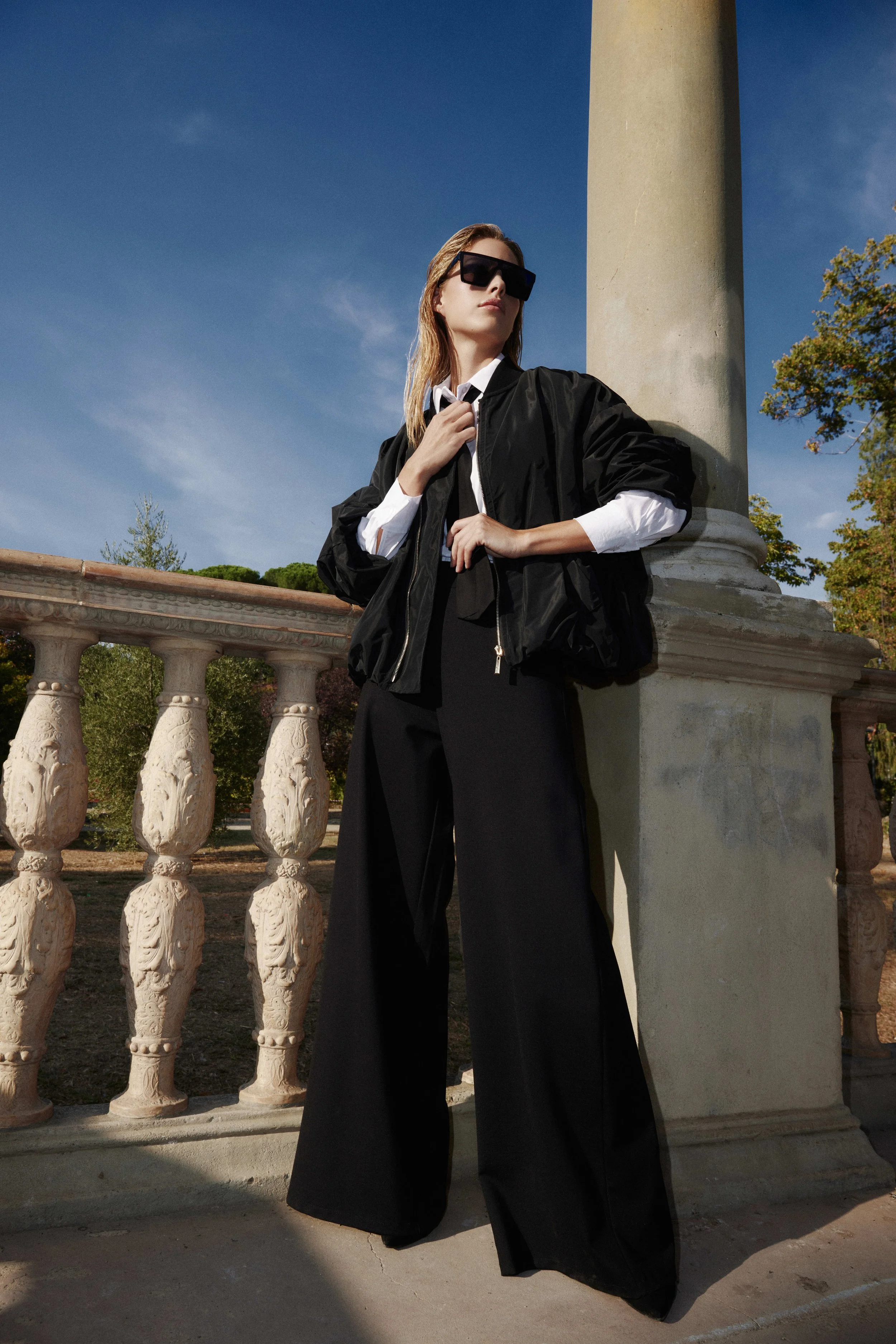 A woman wearing a black jacket, white shirt, black wide-leg pants, and sunglasses, leaning against a stone column outdoors on a sunny day.