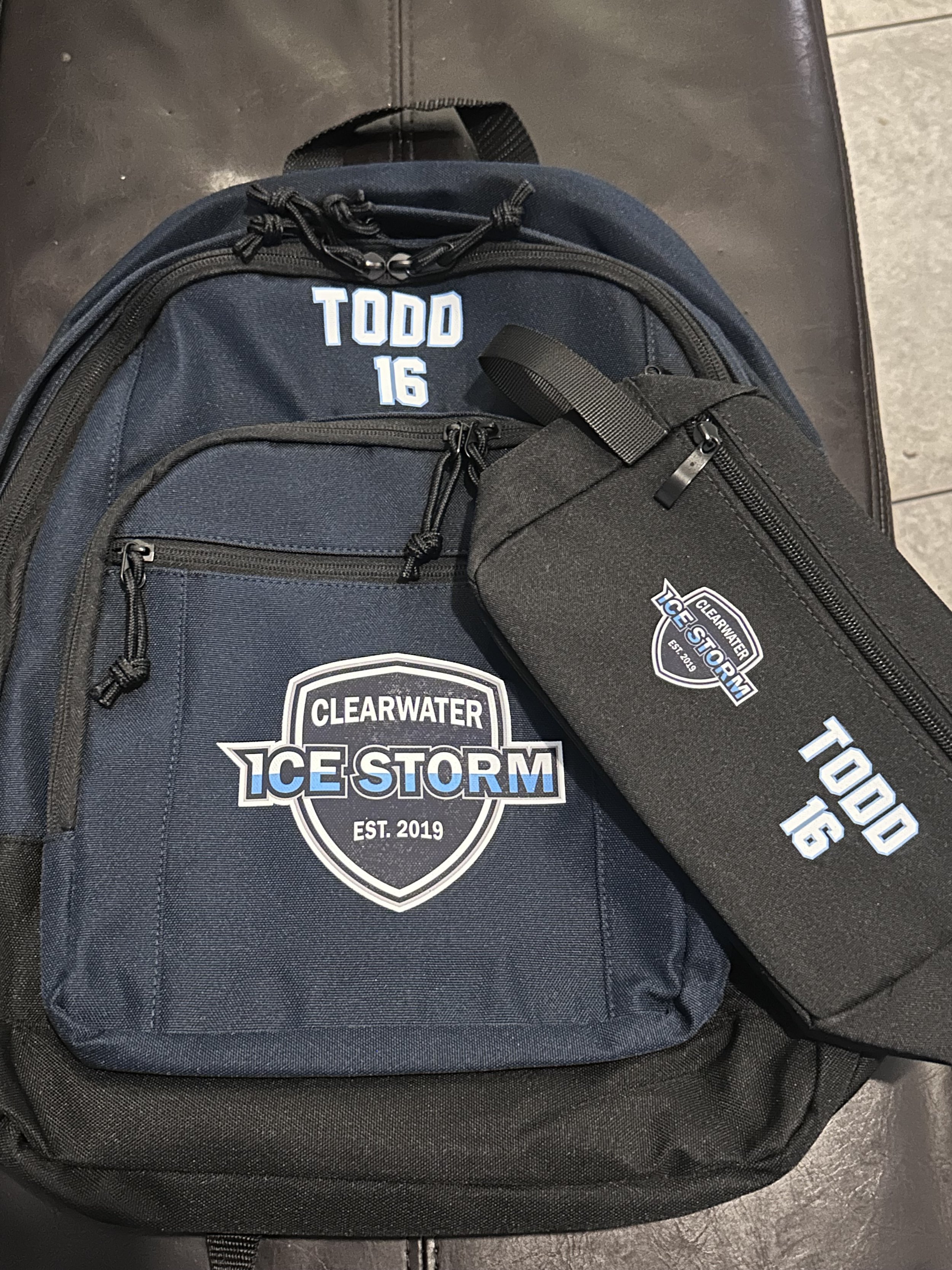 A black backpack with multiple zippered compartments, featuring a white and blue logo of 'Clearwater Ice Storm' and the text 'TODD 16' embroidered on it.