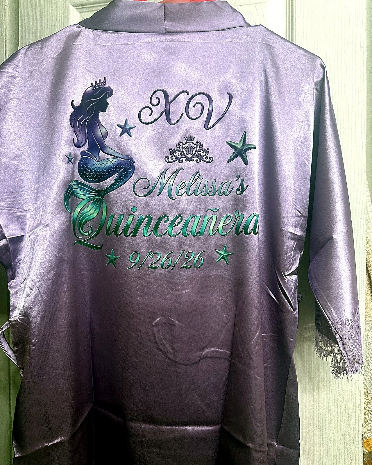 A purple satin satin robe with an embroidered design of a mermaid with flowing hair and a crown, surrounded by stars, and text that reads "XV Melissa's Quinceañera 9/26/26."