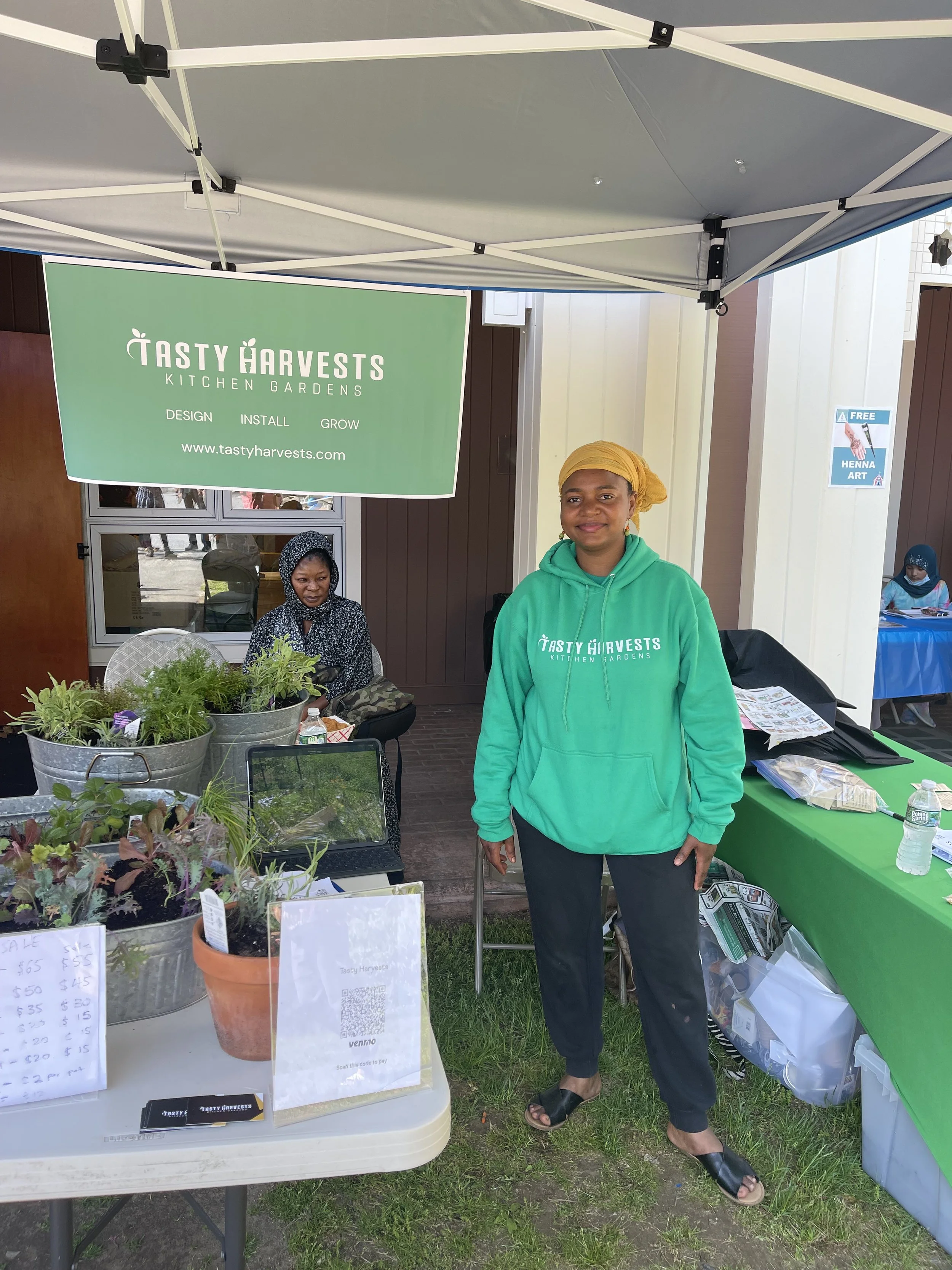 A woman standing at a booth for Tasty Harvests Kitchen Gardens, wearing a green hoodie and a yellow headscarf. The booth displays potted herbs and plants, with a sign overhead promoting design, installation, and growth services for kitchen gardens.