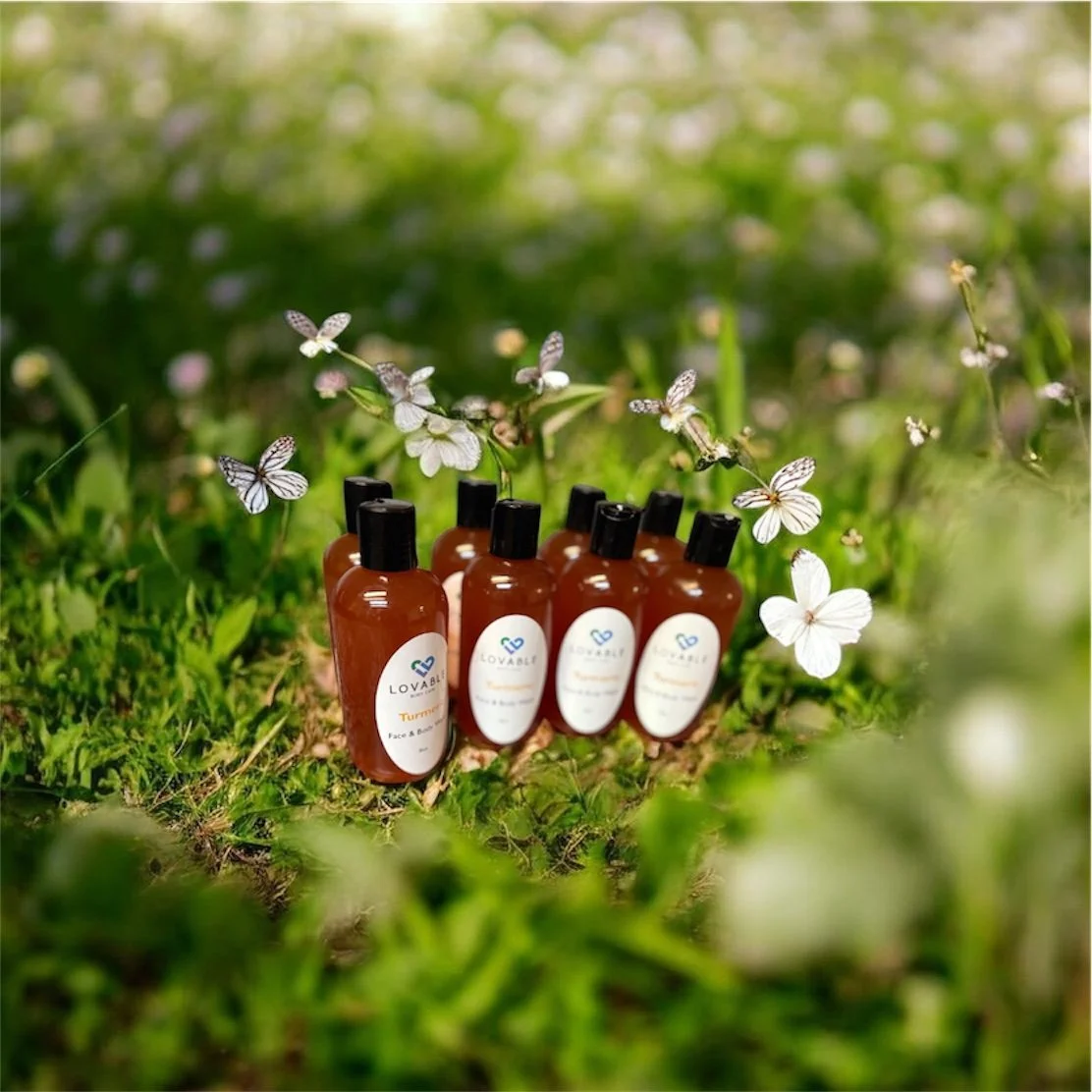 Six brown bottles of Lovable skincare products, labeled "Turmeric Face & Body" and "Love" in a natural green outdoor setting with small white flowers and butterflies.