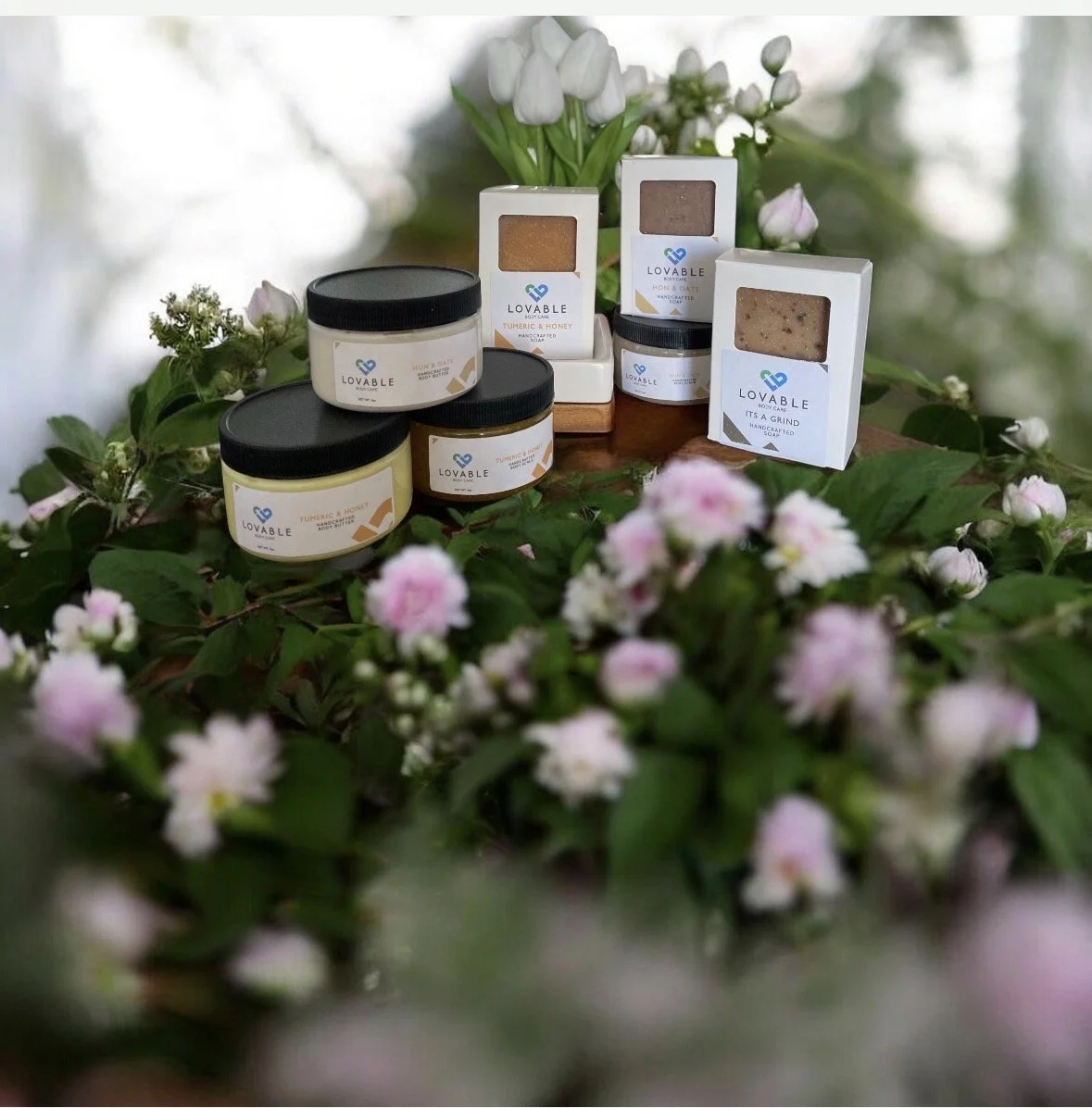 Assorted Lovable body care products, including jars of cream and boxes of soap, arranged on a wooden surface surrounded by pink and white flowers.