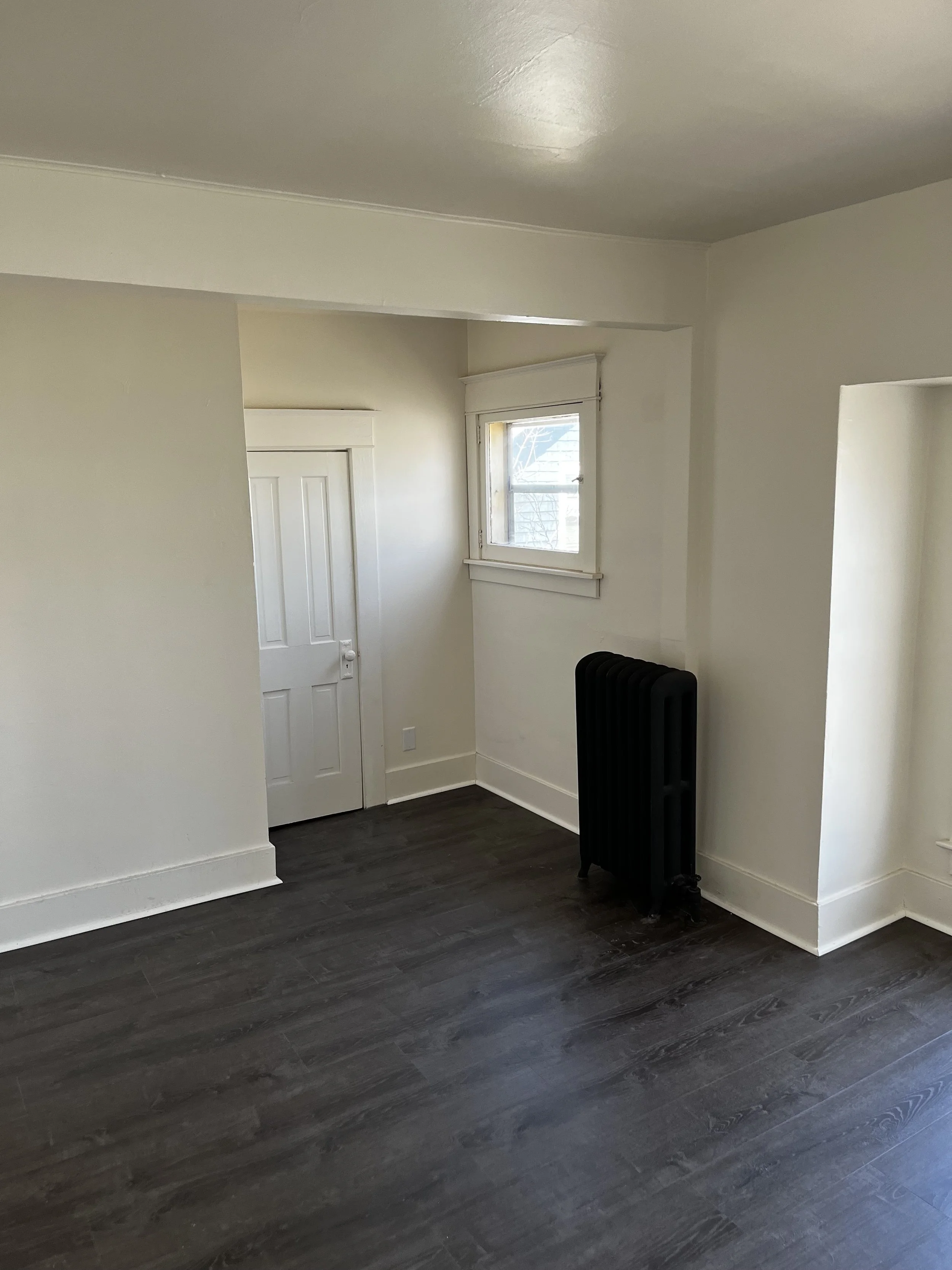 Empty room with white walls, dark wood flooring, a small window, a white door, and a black radiator.
