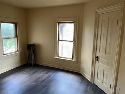 Empty room with two windows, a door, and a radiator, with beige walls and dark flooring.