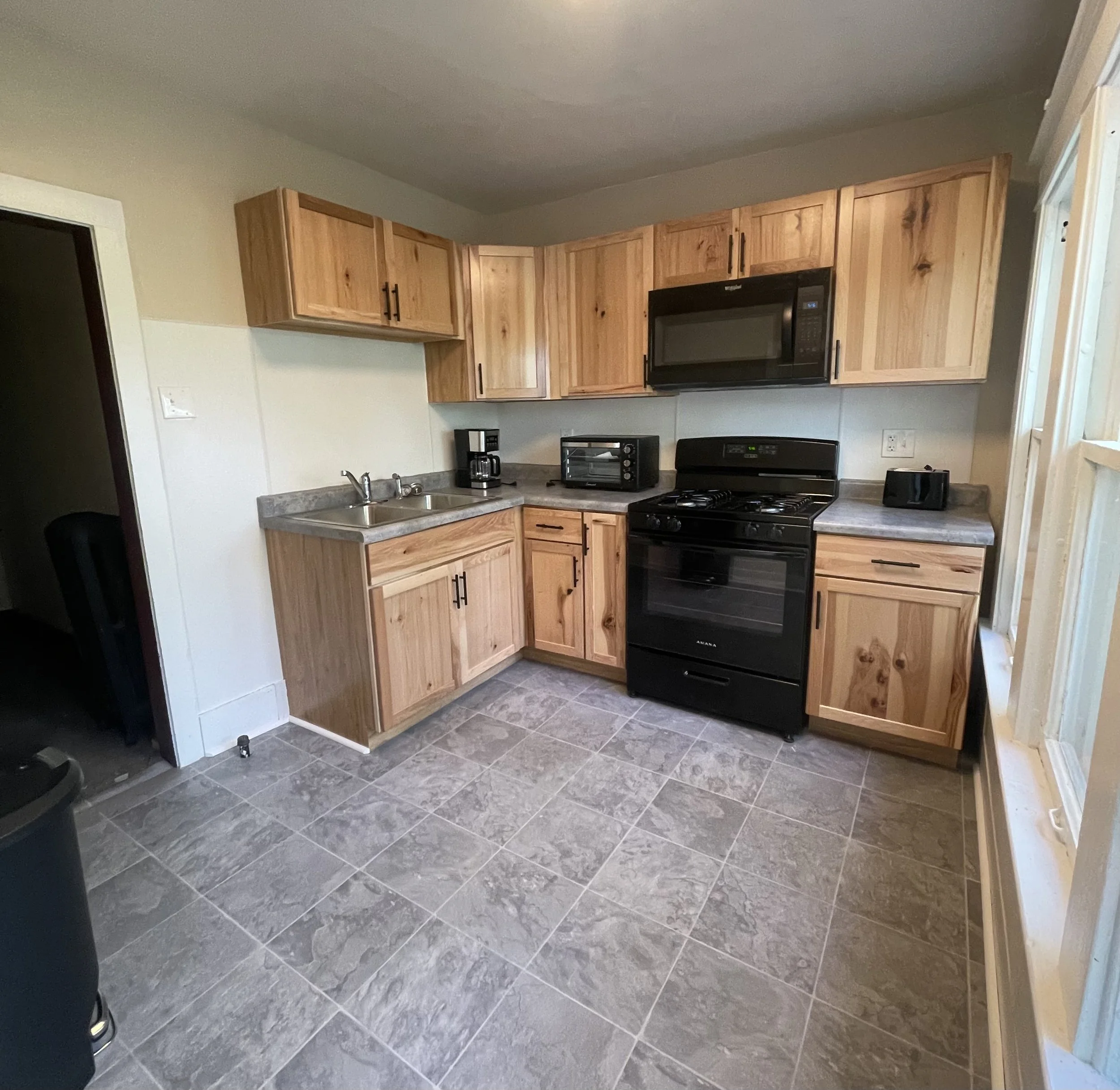 A kitchen with wooden cabinets, a black stove, microwave, toaster, coffee maker, and black countertop. There is a window on the right side and tiled flooring.