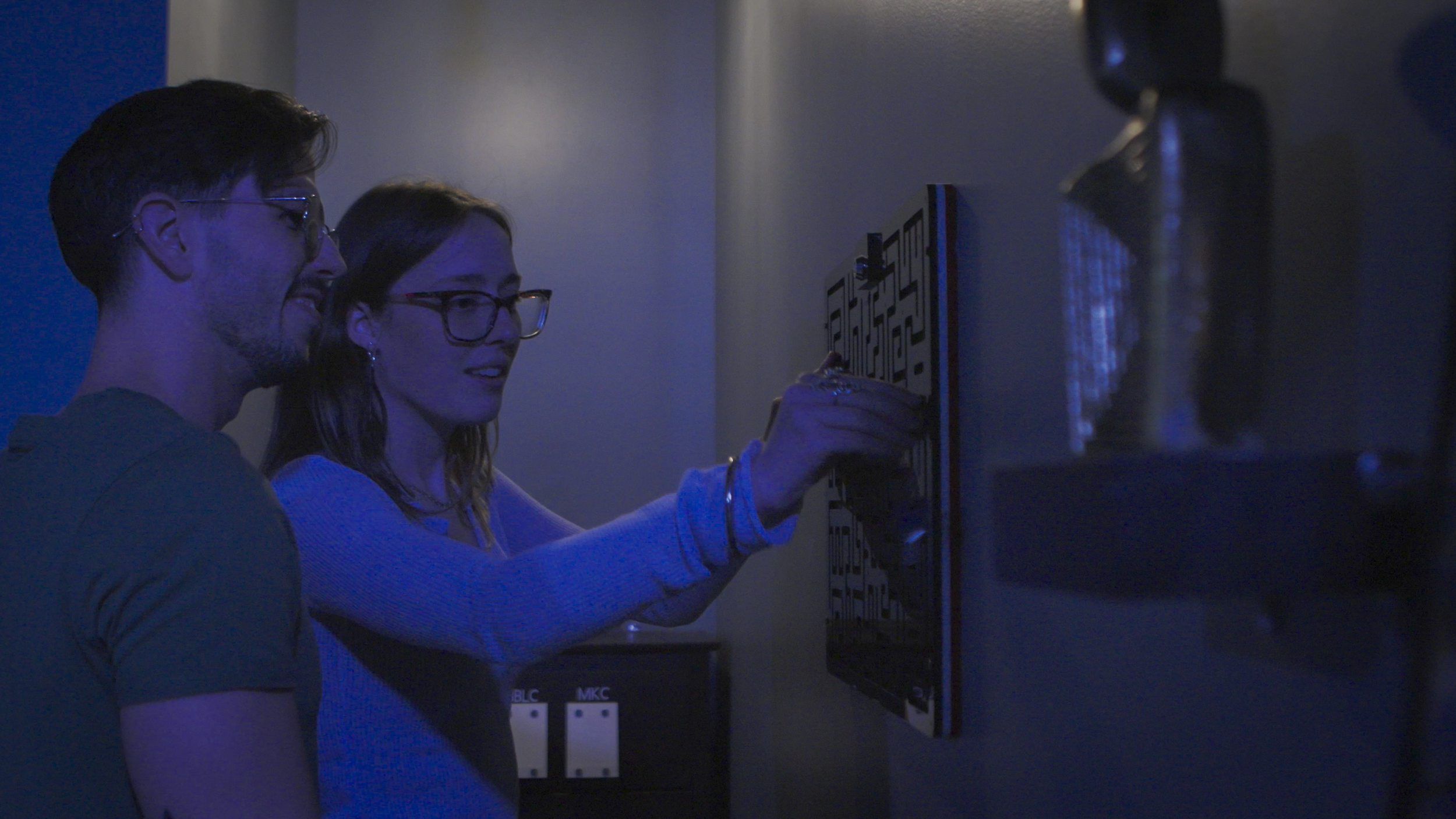 A young man and woman look at a maze artwork on a wall in a dimly lit room with blue lighting.