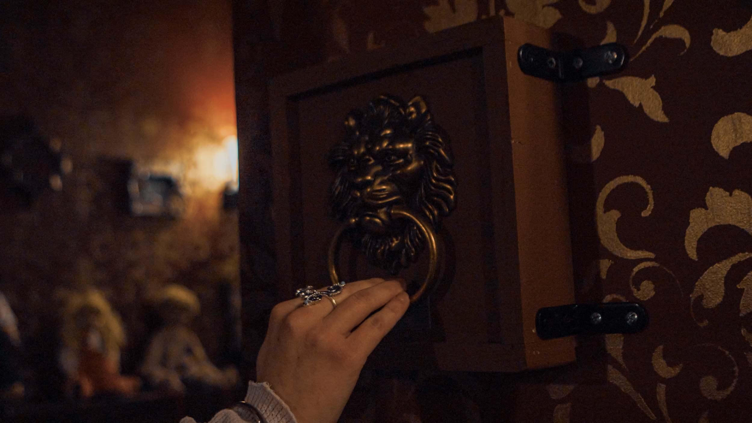 A hand with rings is holding a door knocker shaped like a lion's head on a wooden door.