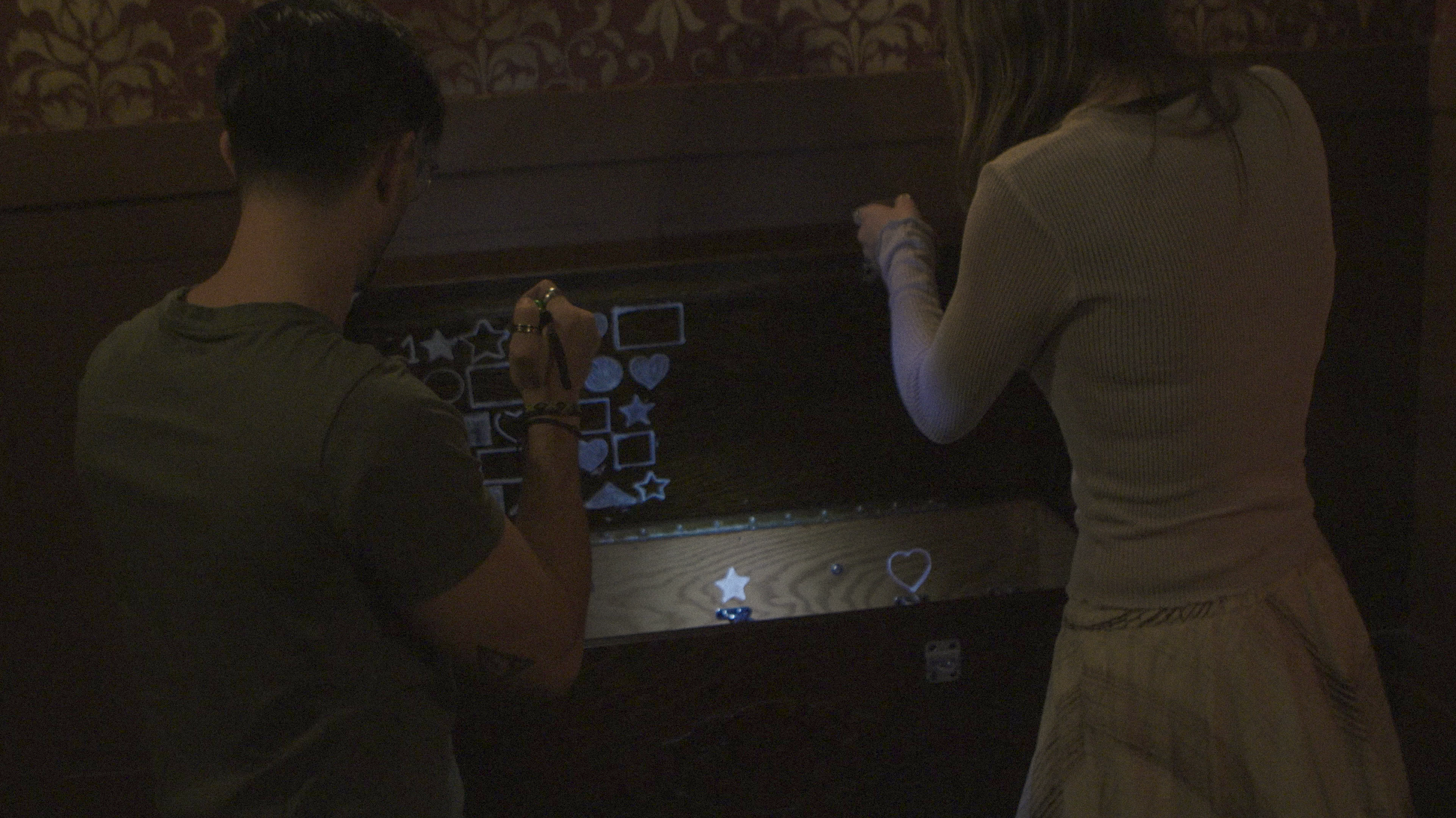 Two people with their backs facing the camera are drawing with light on a dark wooden surface. The person on the left is sitting and wearing a short-sleeved shirt, while the person on the right is standing and wearing a long-sleeved top and a skirt. 