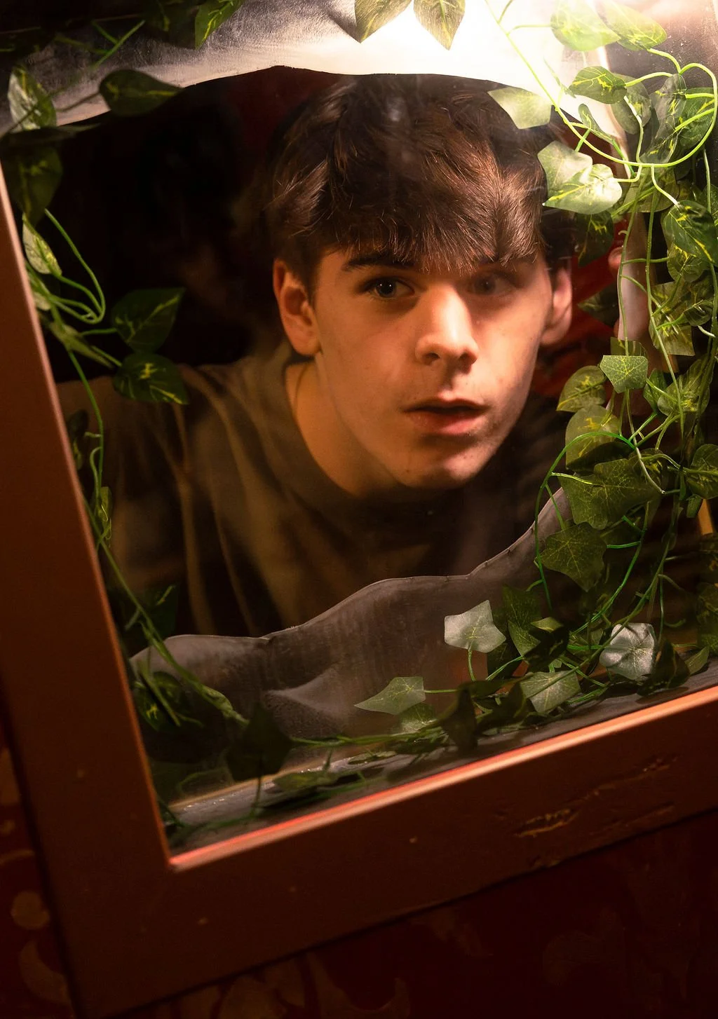 A young man with brown hair and a surprised expression looking into a glass display case filled with green leafy vines.