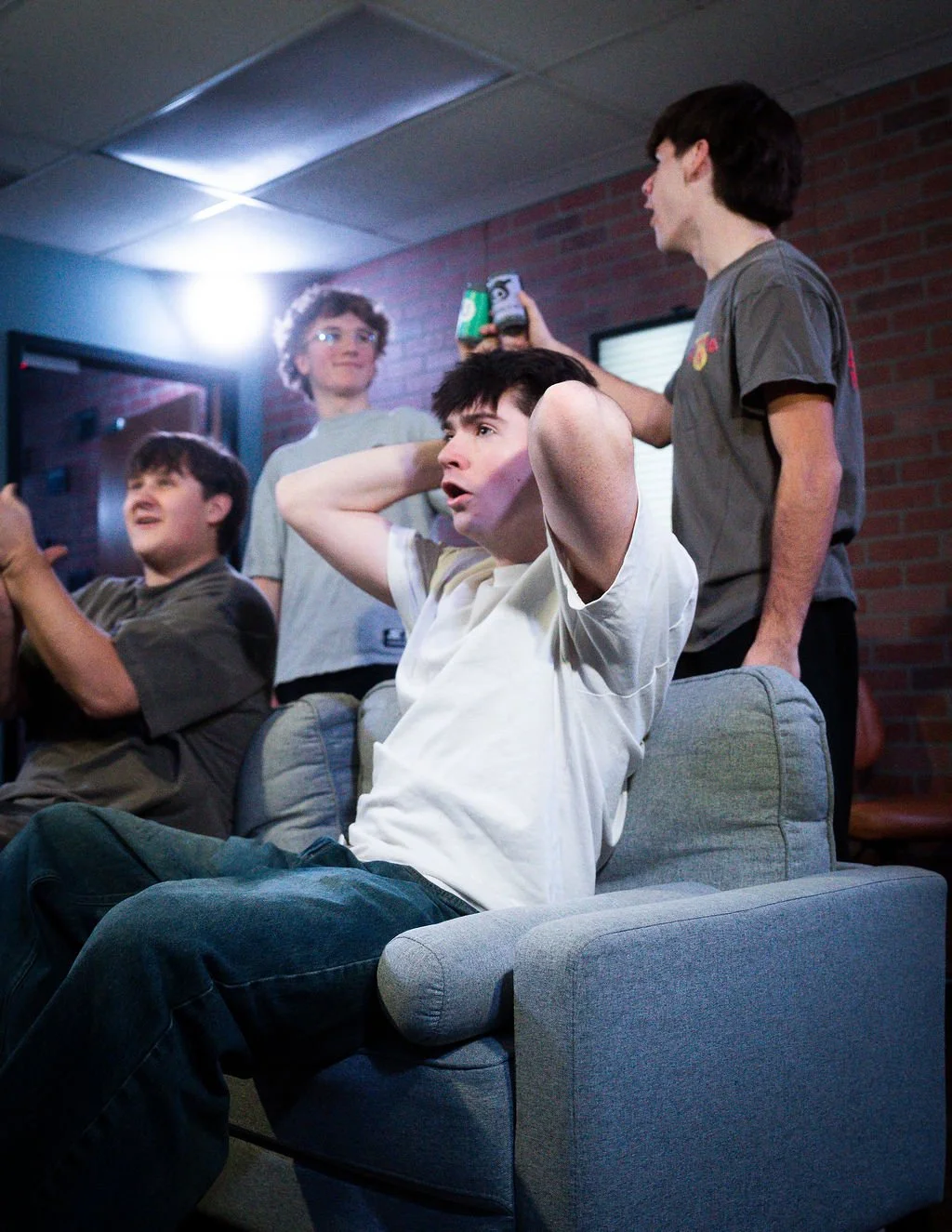 A group of young men gathered in a casual setting, with one seated on a couch holding his head in surprise or shock while others are standing and interacting.