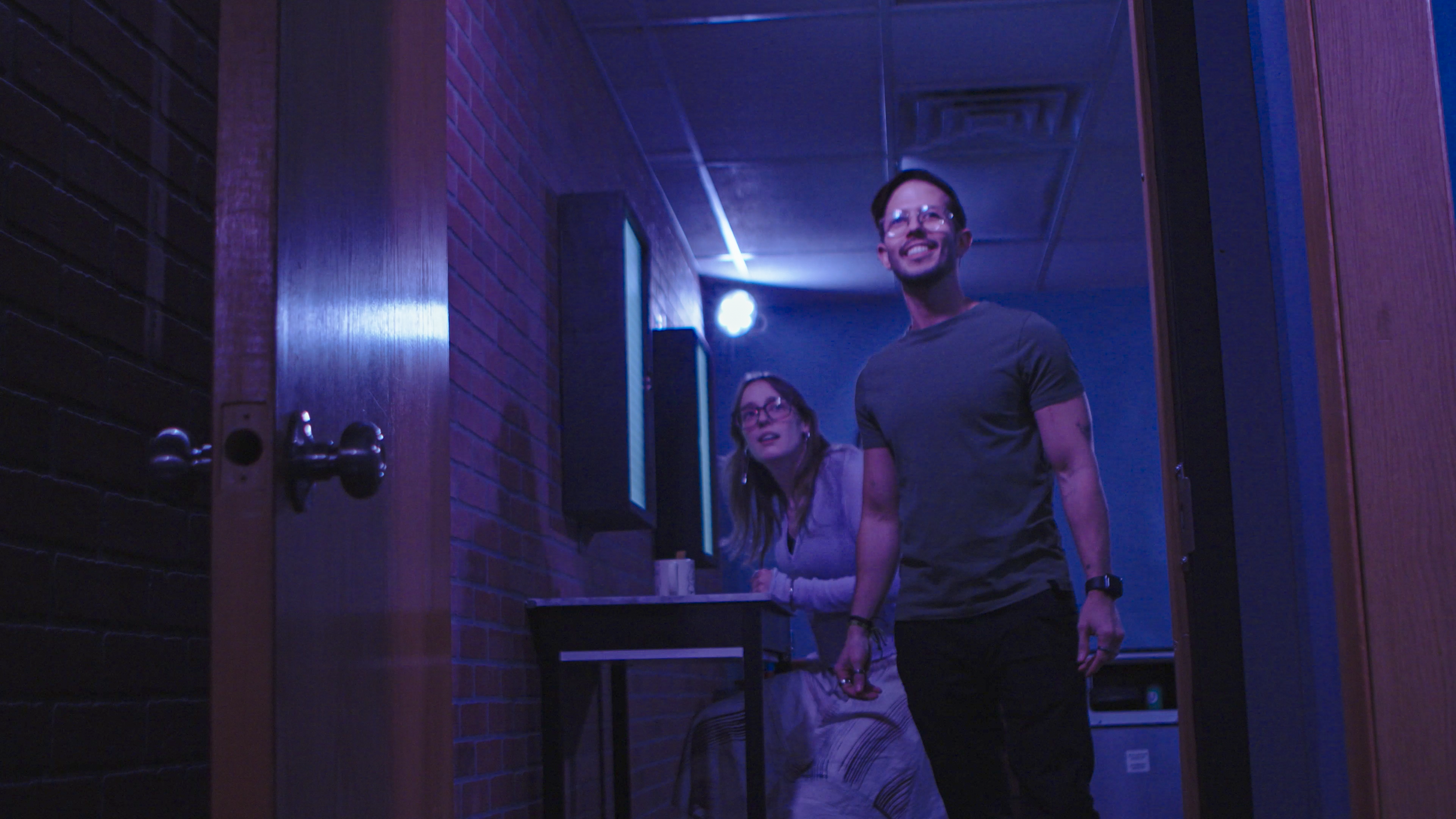 Two people, a man and a woman, in a karaoke room with blue and purple lighting. The man is smiling and facing the camera, while the woman looks surprised or curious.