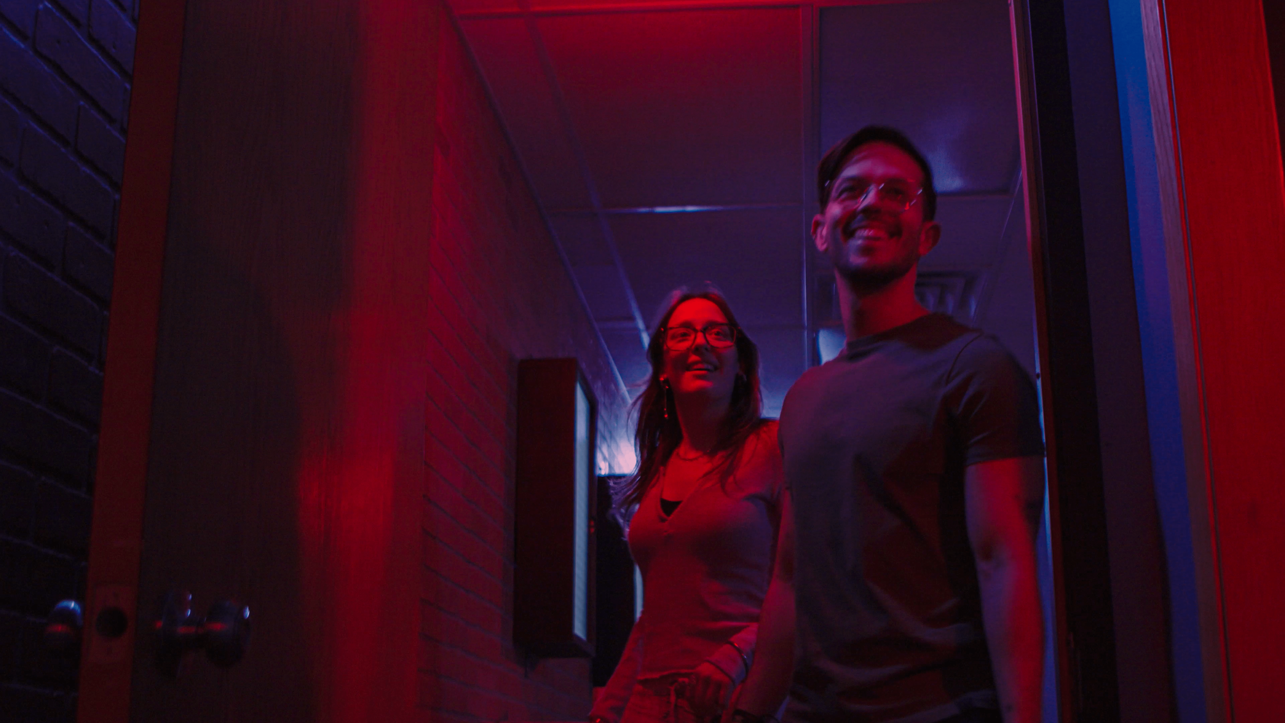 Two smiling people standing in a doorway illuminated by red and blue neon lights in a dimly lit room.