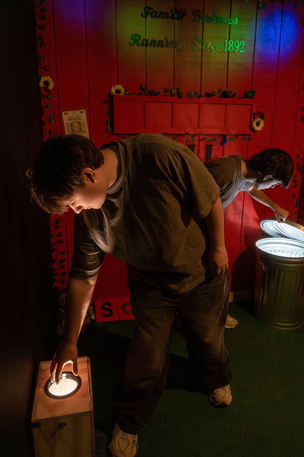 Two young men playing a game in a dimly lit room with red walls decorated with flowers and green lettering that says 'Family Funplex Runningsince 1892'.