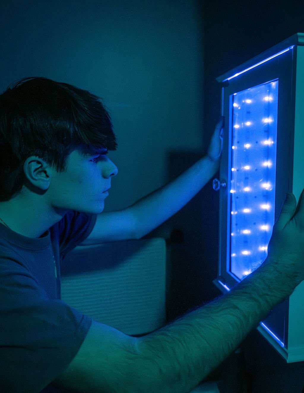 A young person with dark hair interacts with a small, illuminated LED light box in a dark room.