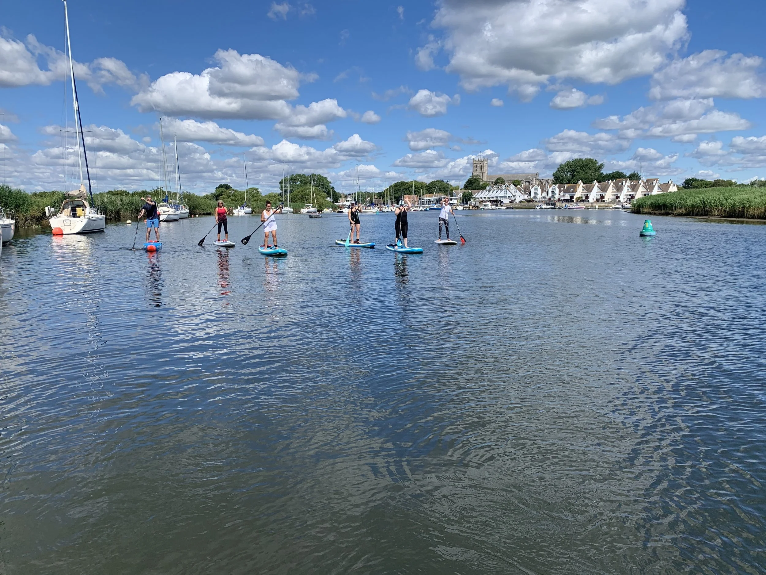 People paddleboarding on a calm water canal under a blue sky with scattered clouds, with boats and houses visible in the background.
