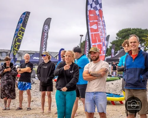 Group of people gathered outdoors near the beach, with flags and tents in the background, some smiling and talking.