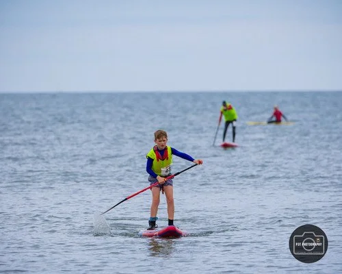 People paddleboarding in the ocean with a young person in the foreground wearing a yellow vest and blue shirt.