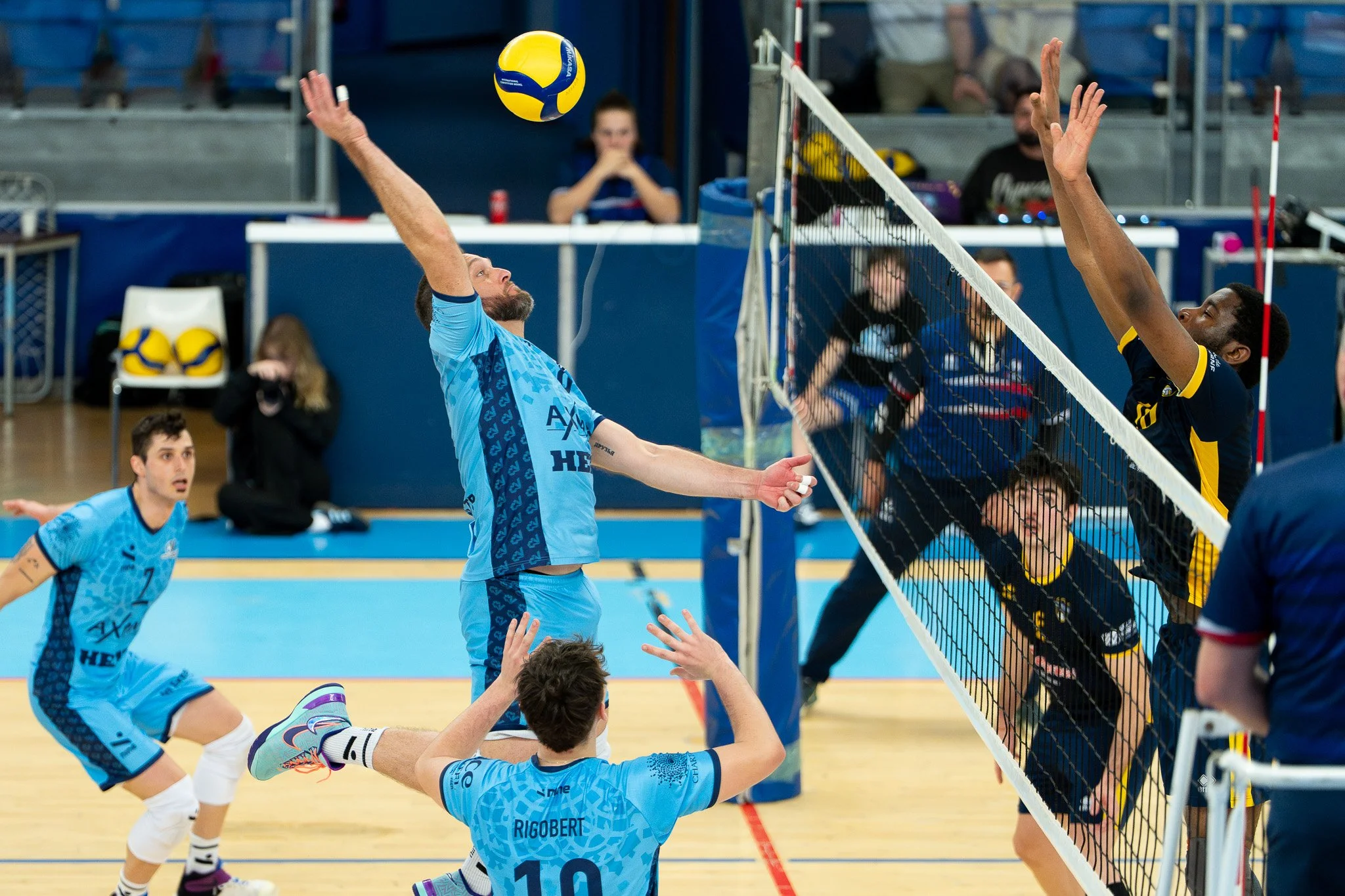 Joueurs de volleyball en action lors d'un match, avec un joueur en bleu sautant pour frapper le ballon au-dessus du filet contre l'équipe en noir et jaune. C'Chartres Volley