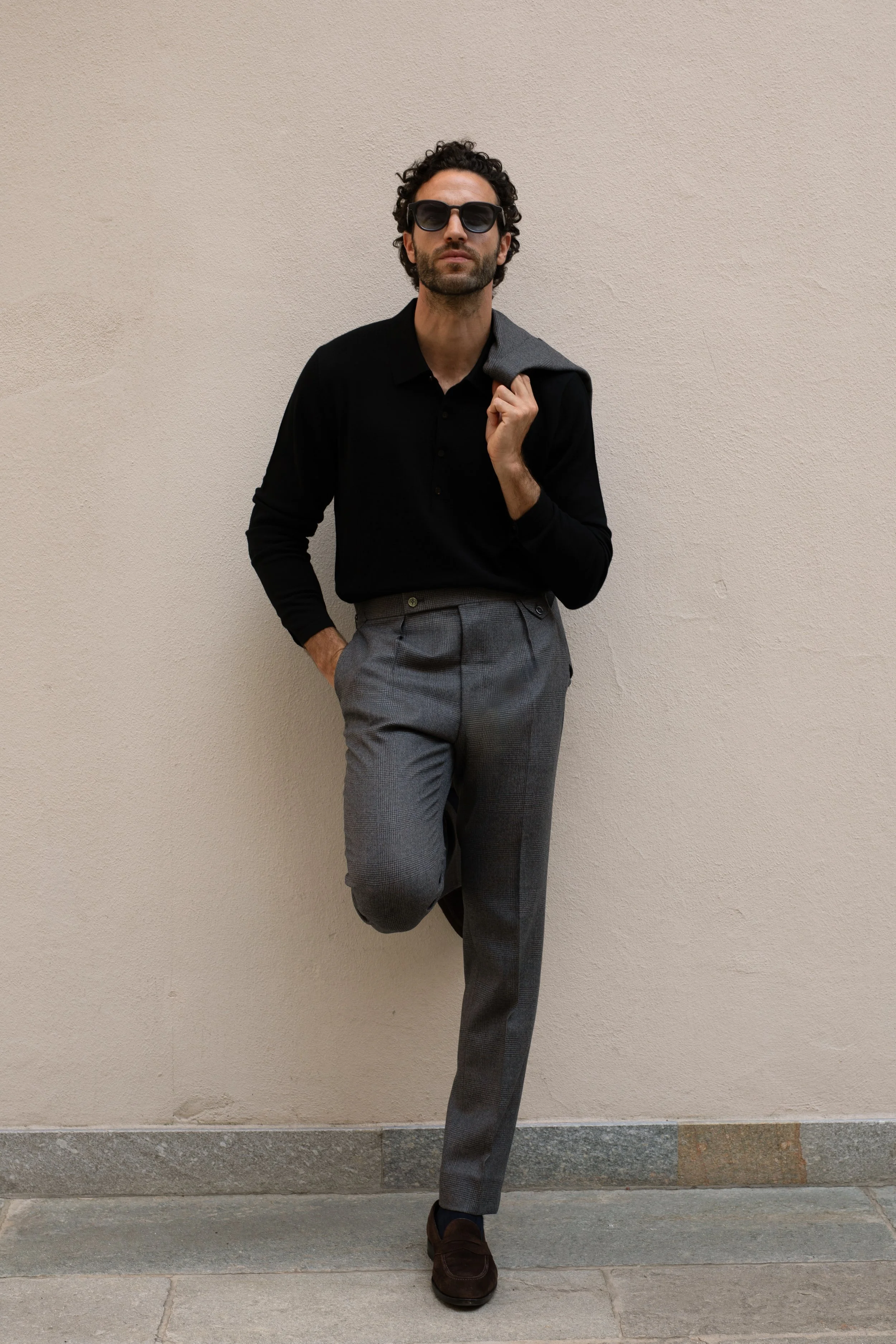 A man wearing sunglasses, a black long-sleeve polo shirt, gray checkered pants, and dark shoes, standing against a beige wall with his left hand in his pocket and his right hand holding a gray jacket over his shoulder.