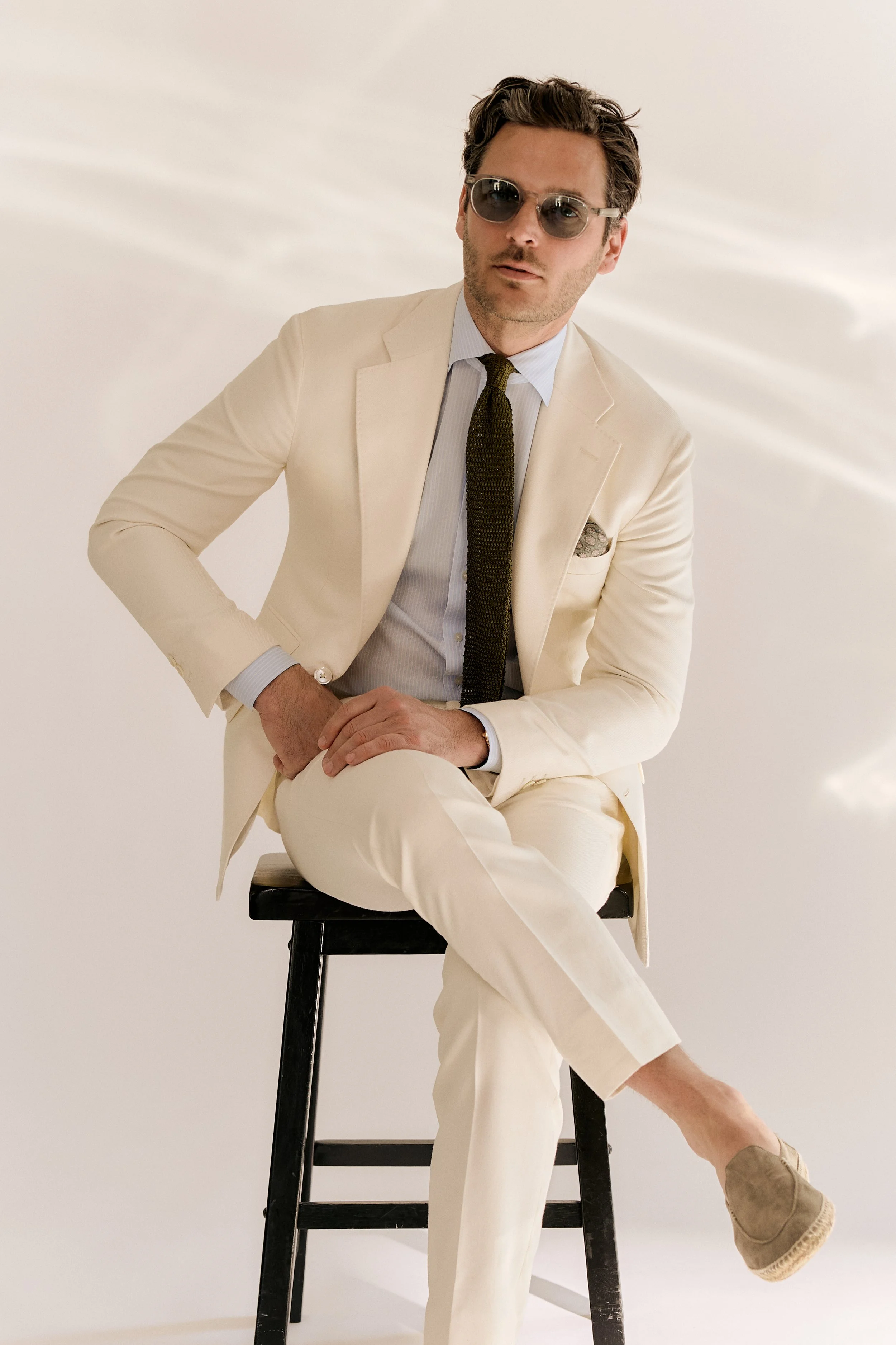 A man with dark hair, wearing sunglasses, a beige suit, white shirt, and dark tie, sitting on a black stool against a white background.