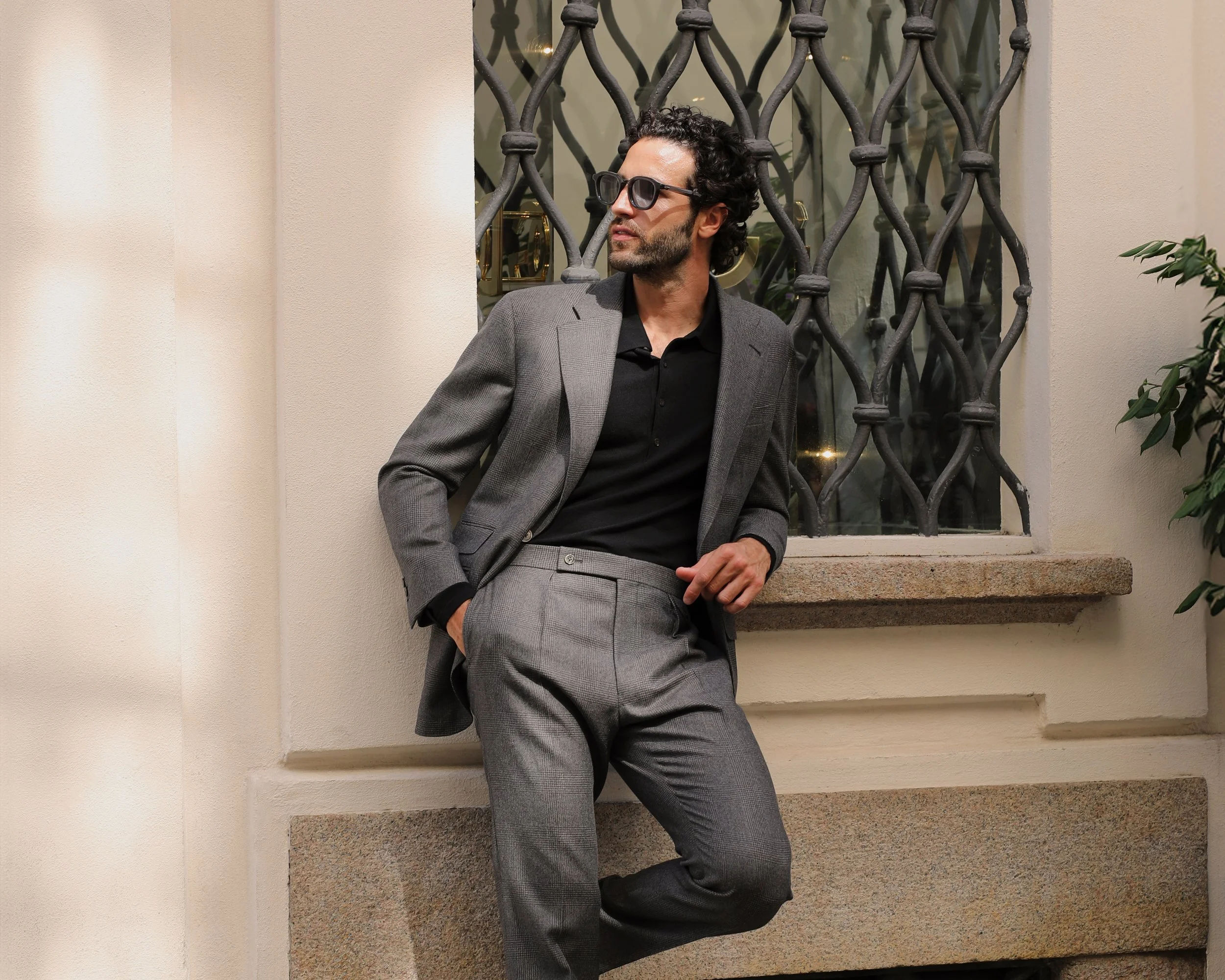 A man with curly dark hair, beard, and wearing sunglasses leaning against a beige wall with a window behind him. He is dressed in a gray suit with a black shirt, with his left hand in his pocket and right hand resting on his belt.