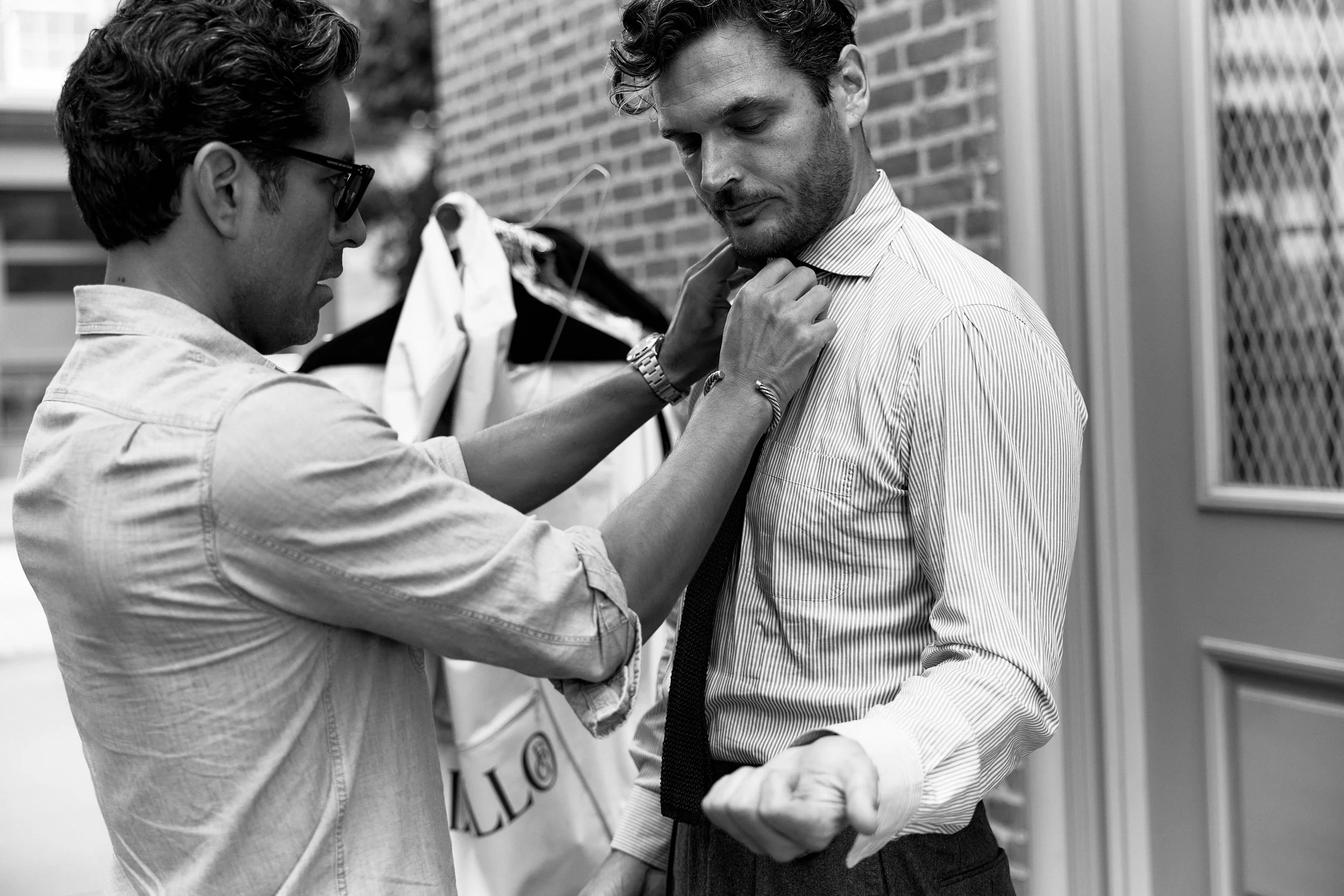 A man in a light-colored shirt adjusts his tie as another man, wearing glasses and a watch, helps him by adjusting his collar outside against a brick wall.
