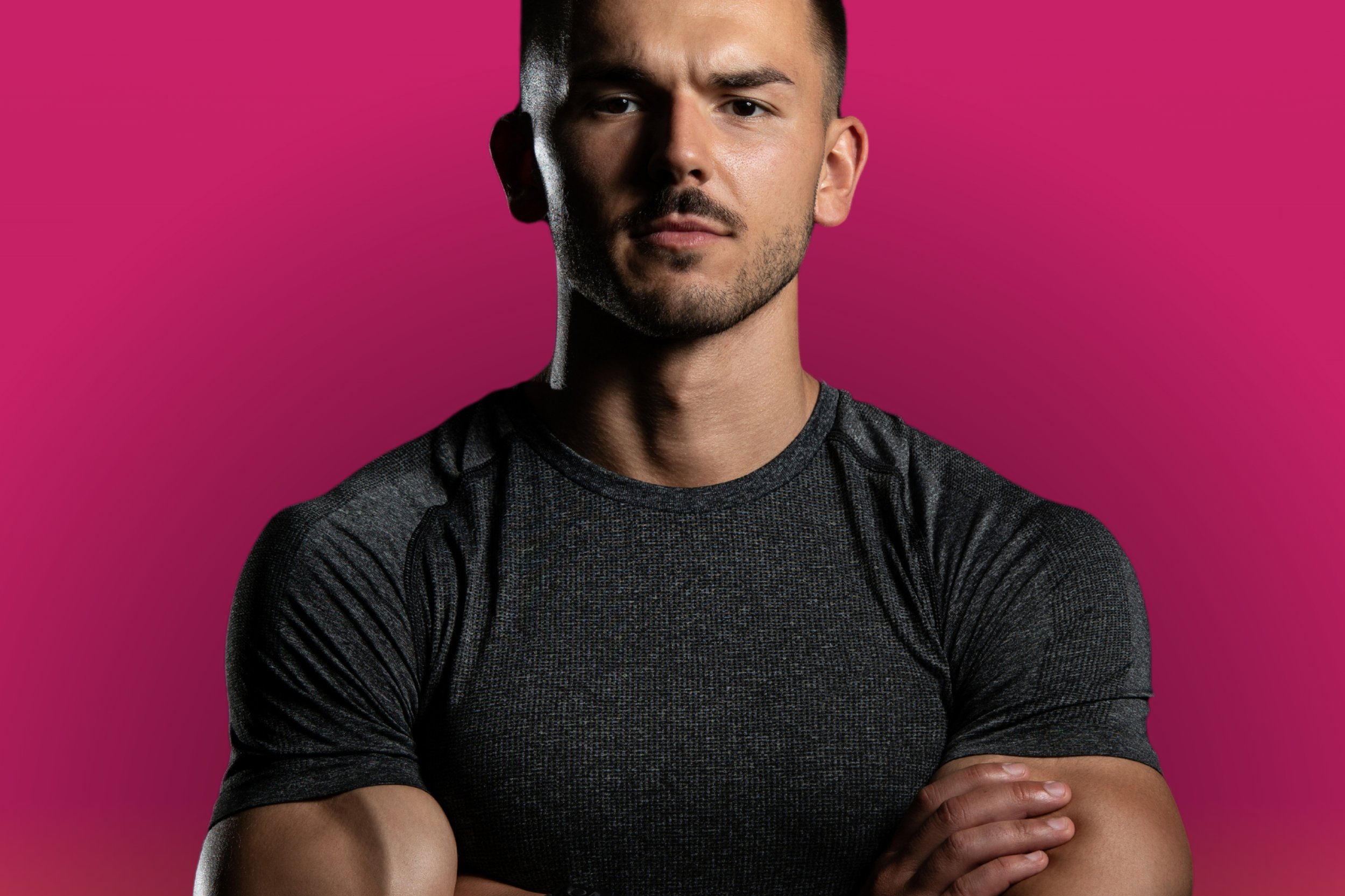 Man in gray athletic shirt with arms crossed against a pink background.