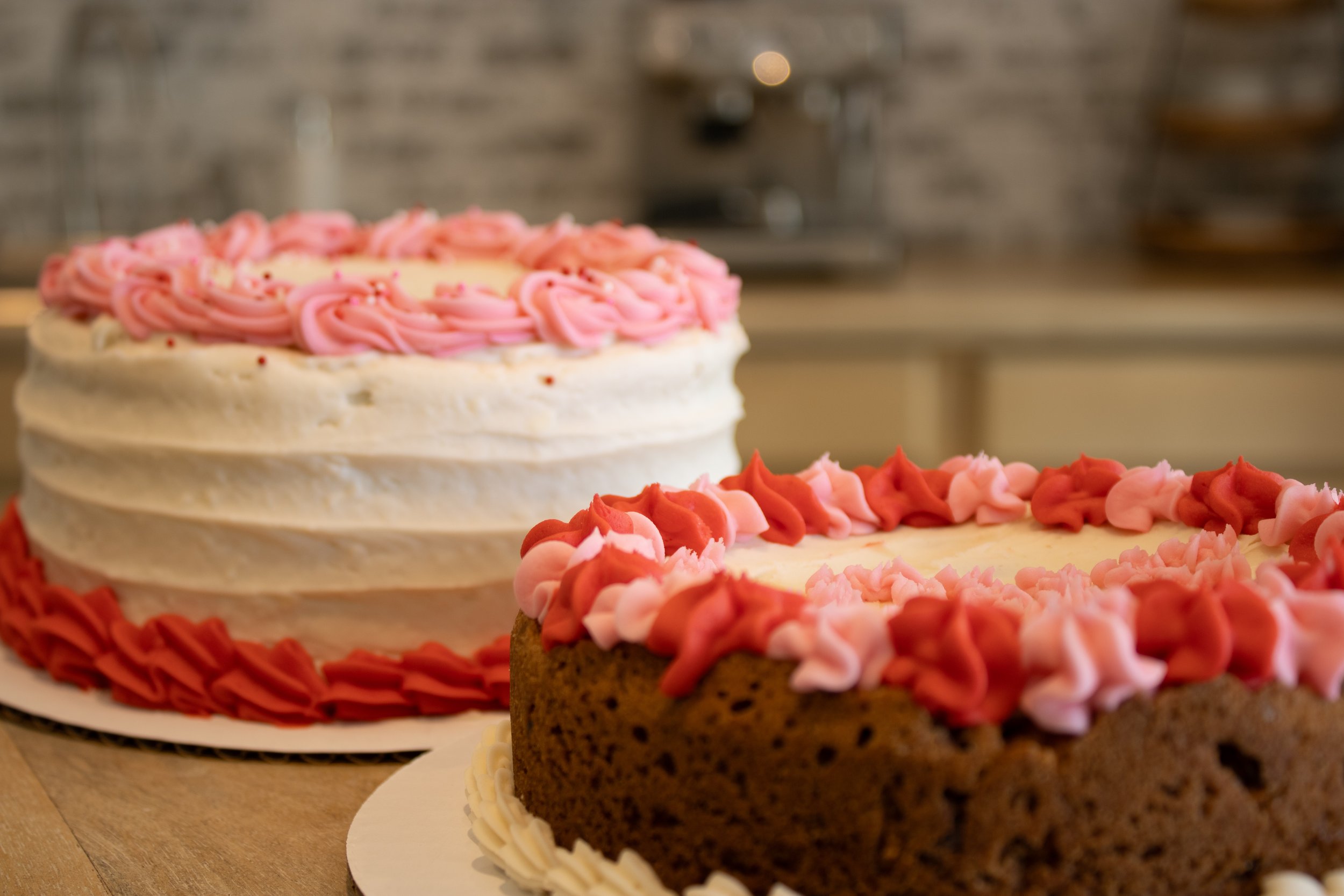 Two decorated cakes on a wooden table with a brick wall background, one with white icing and pink piping, and the other with chocolate base and pink and red piping.