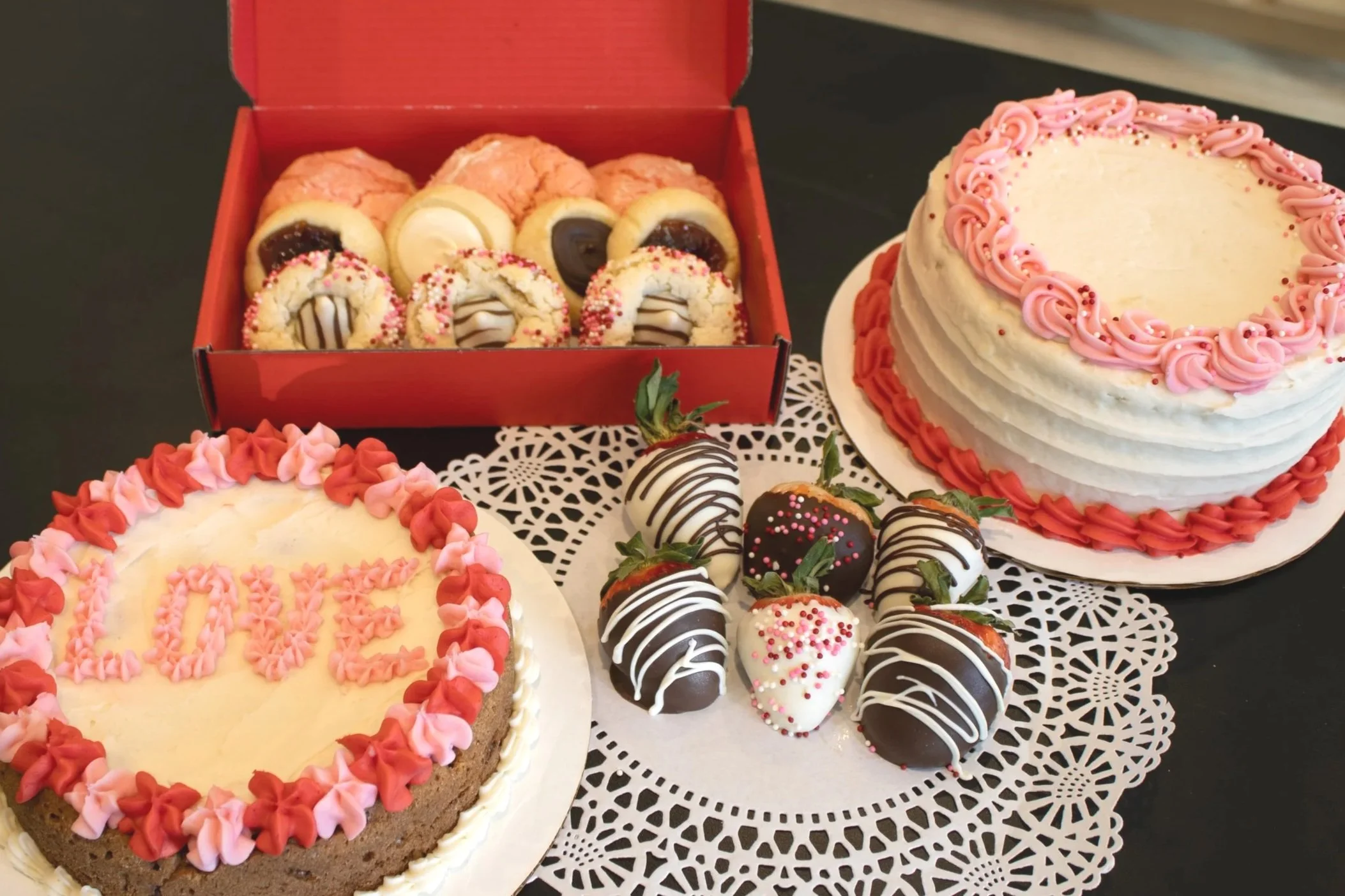 A variety of desserts on a table including two cakes with pink and red icing, a platter of chocolate-dipped strawberries, and a box of assorted cookies and pastries.