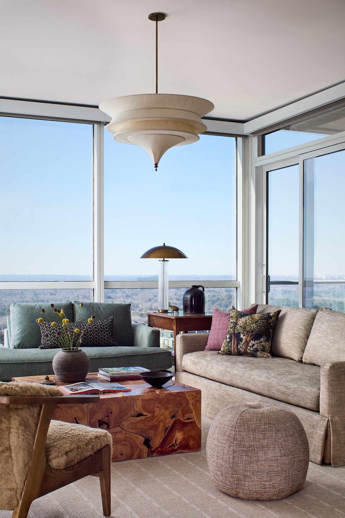 high rise living room with floor to ceiling windows, seating is anchored by a statement pendant lighting, organic wooden coffee table and city views