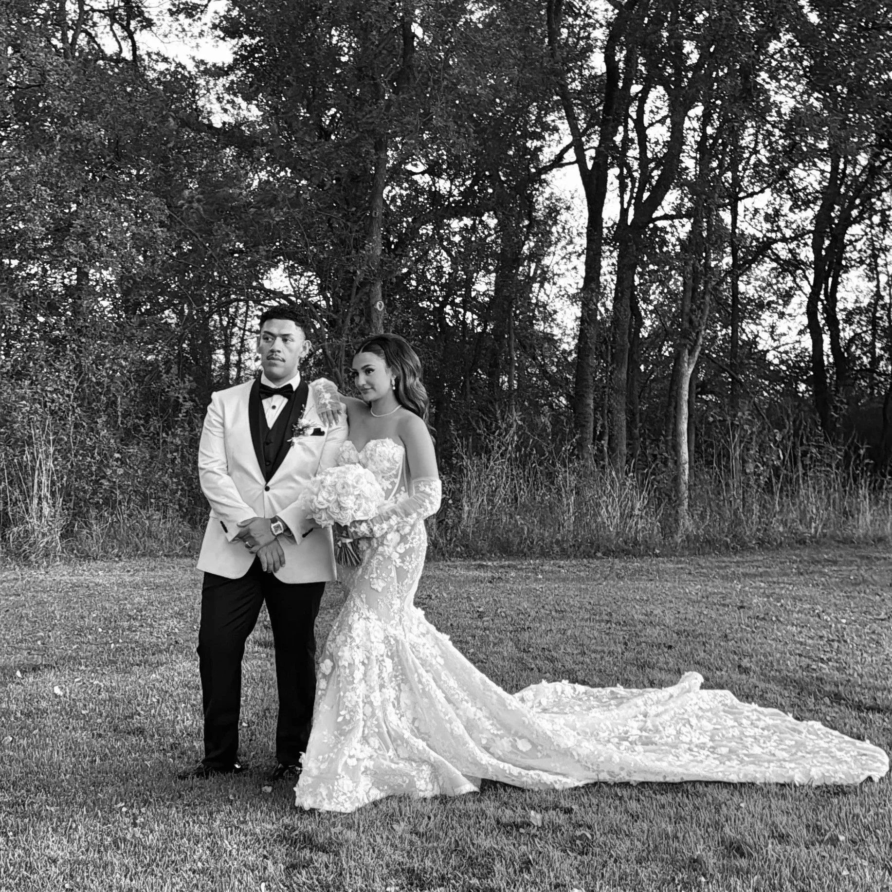 A bride and groom standing close together outside a white building at sunset, with the bride in a long white wedding dress and the groom in a black suit and cowboy hat.