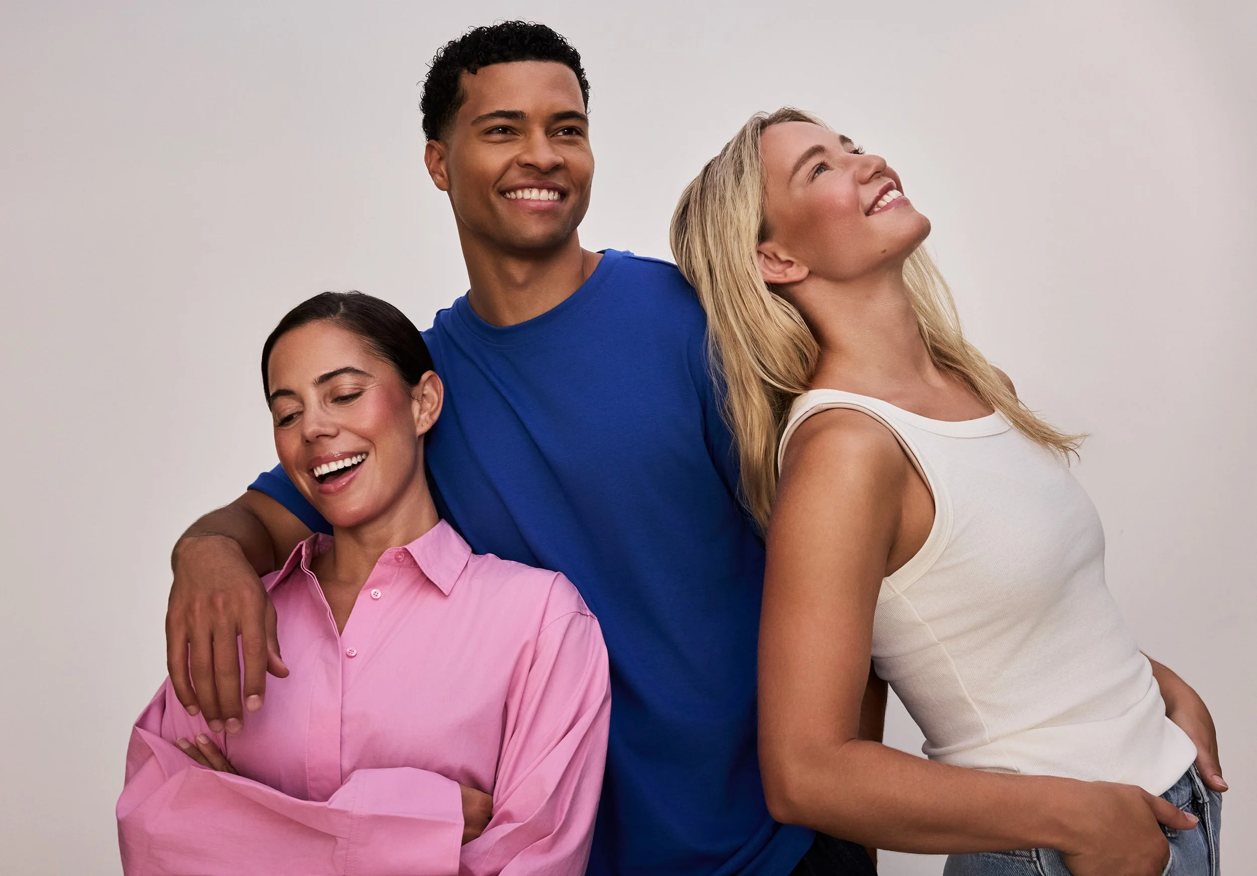 Drei lachende Menschen, zwei Frauen und ein Mann, die sich gegenseitig umarmen und in eine positive Richtung schauen, vor einem neutralen Hintergrund.