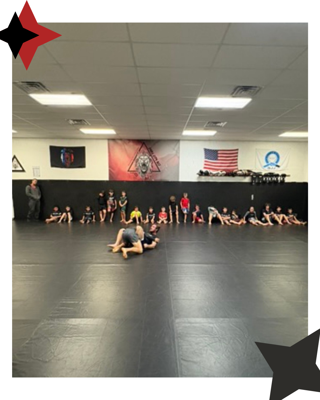picture showing instruction of a kids class with children seated against the wall and professor and coaching demonstrating the technique in front of the class