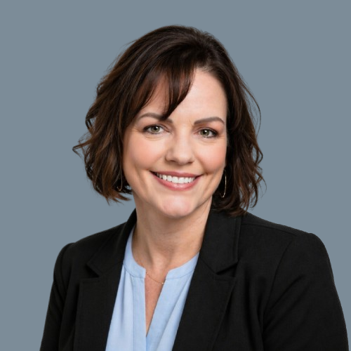 Portrait of a smiling woman with short brown hair, wearing a black blazer and light blue blouse, against a gray background.