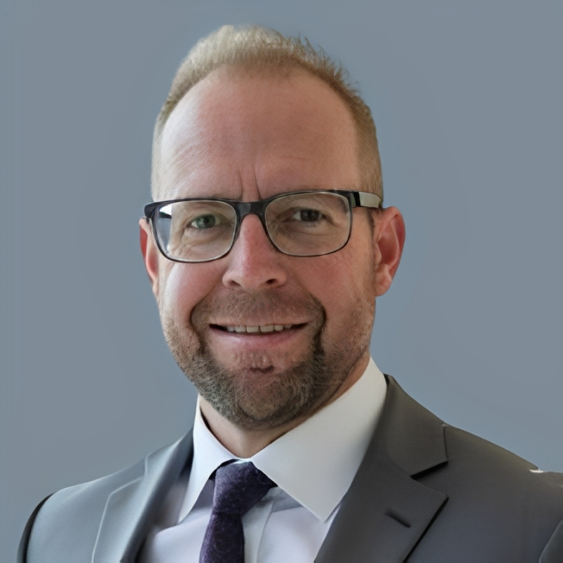 A man wearing glasses, a suit, and a tie, smiling against a plain background.
