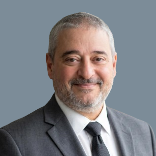 Headshot of a middle-aged man with gray hair and beard, wearing a gray suit and tie, smiling against a plain background.