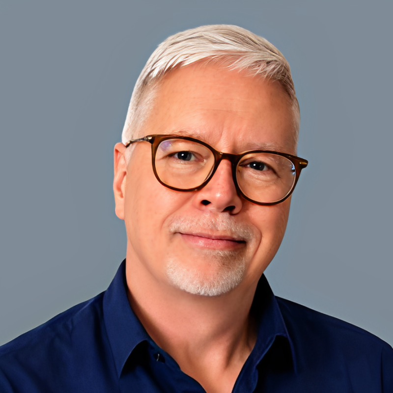 Portrait of a middle-aged man with short gray hair, glasses, and a goatee, wearing a dark blue shirt, smiling softly against a plain gray background.