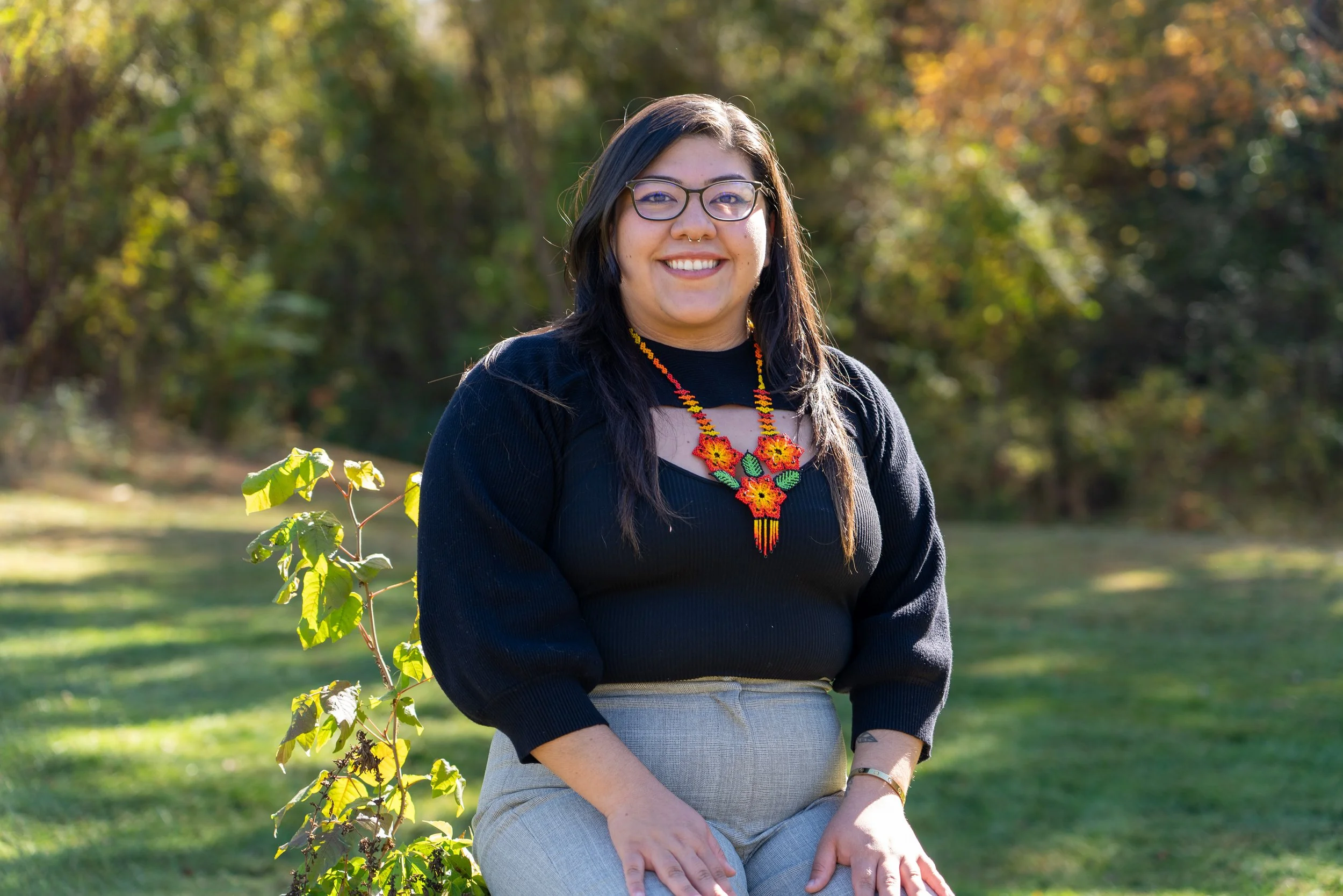 Monse sits while wearing a colorful beaded necklace. She has a black long sleeve with a cutoff.