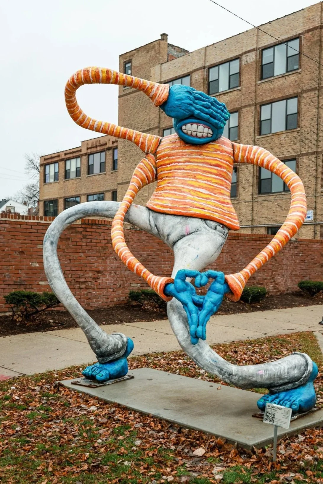 Colorful street art sculpture of a figure with blue hands and feet, orange and white striped limbs, wearing an orange striped shirt and white pants, making a hand heart gesture, in an outdoor urban setting with brick buildings and fallen leaves.