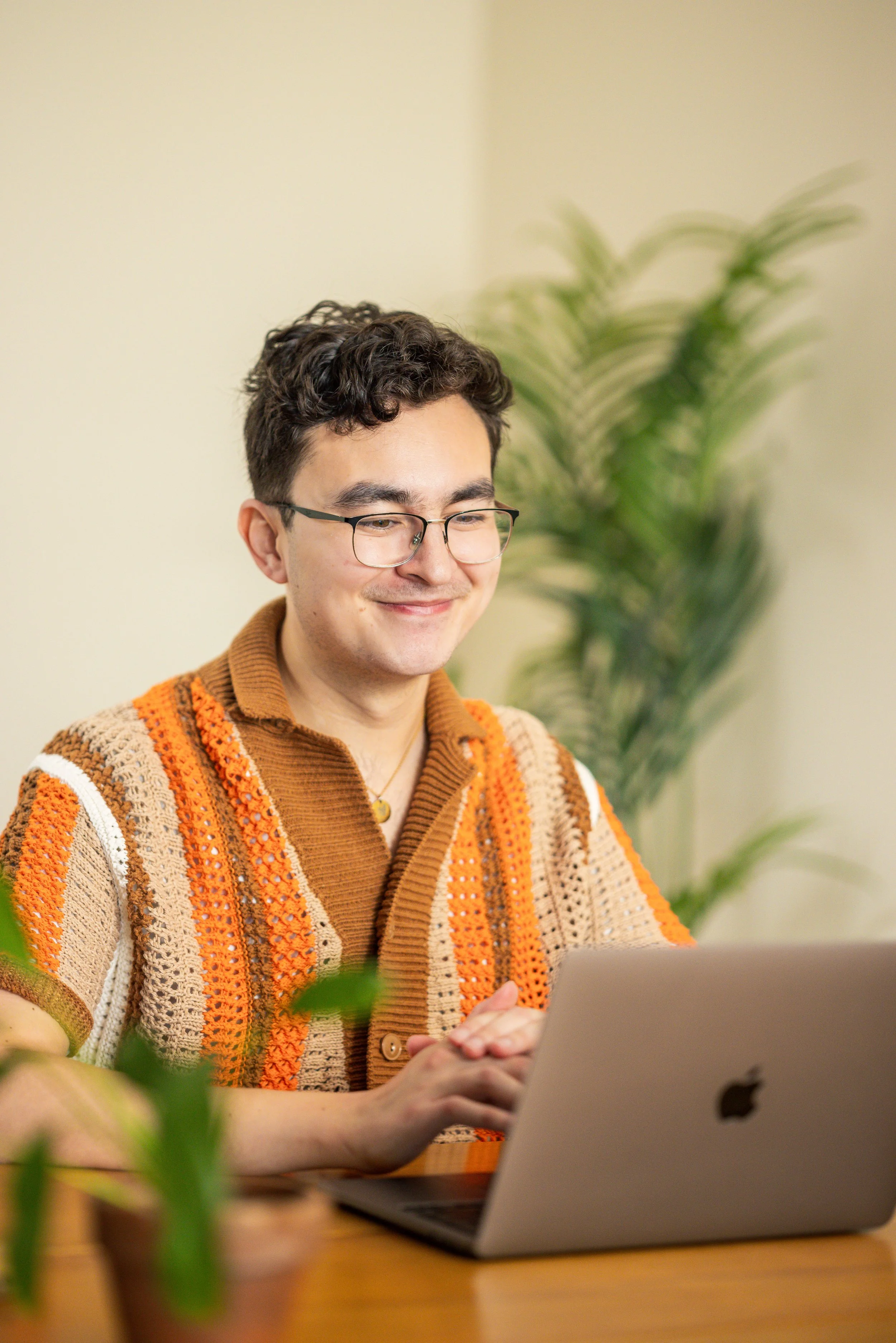 Glasses and dark curly hair, smiling, working on a silver MacBook. He is wearing a colorful knitted sweater with orange, beige, and brown patterns. There are green plants in the background and foreground, and the room has a neutral-colored wall.