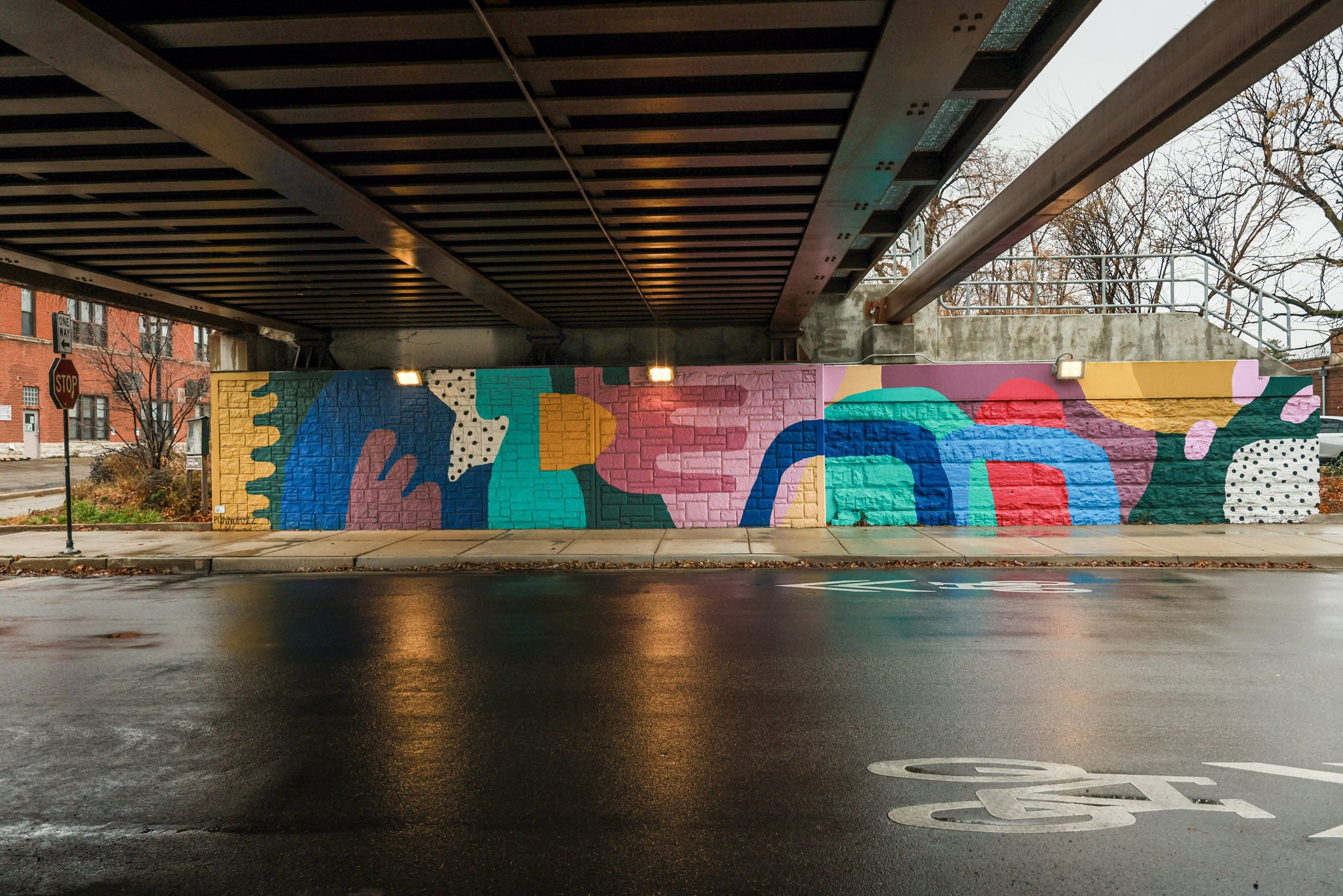 Underpass with colorful abstract mural painted on the brick wall beneath the bridge, with a wet street and a bicycle lane marked on the road.
